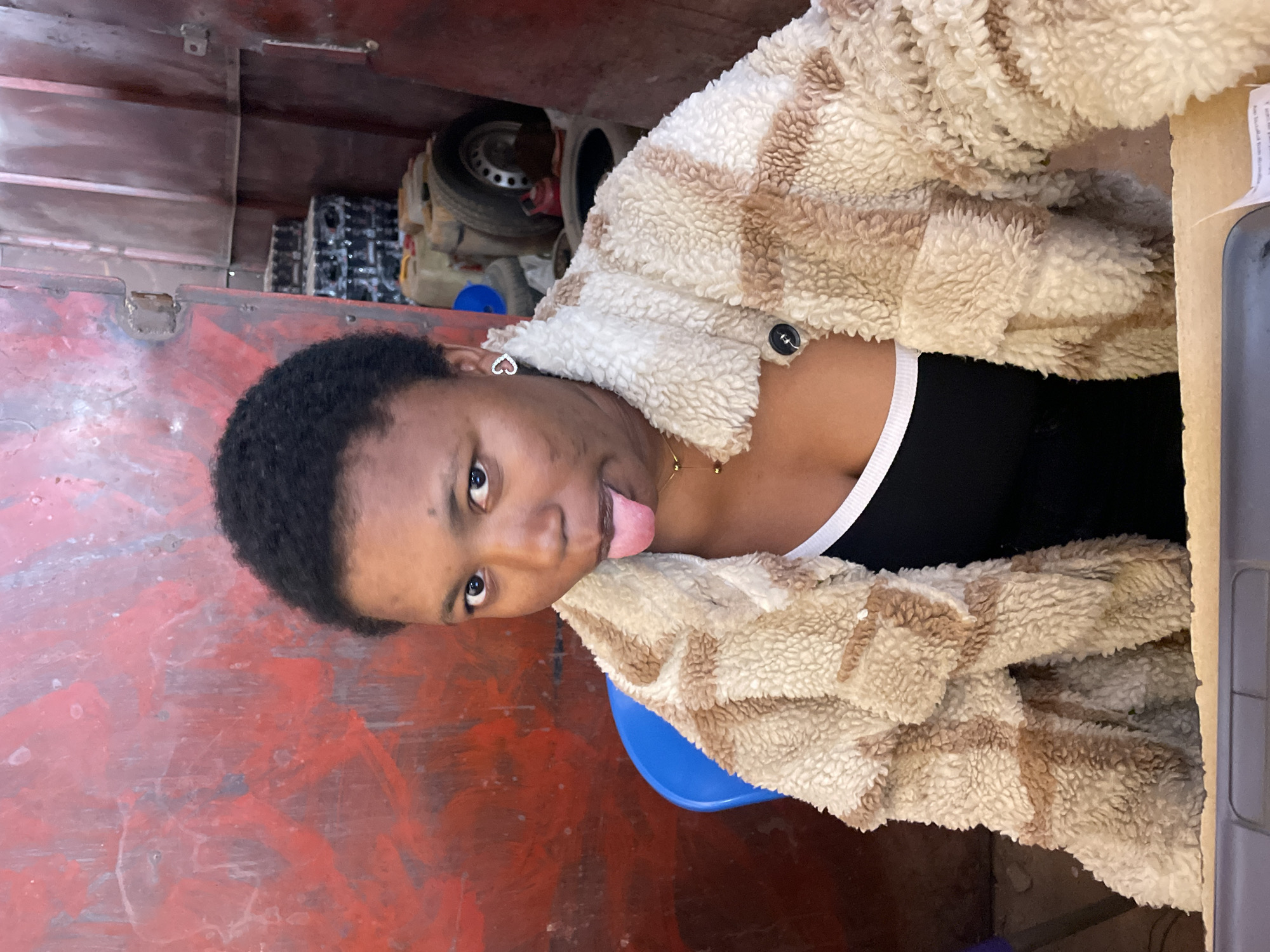 A young, dark-skinned woman with short, dark hair is sticking her tongue out at the camera. She is wearing a black top with a white trim and a fuzzy, patterned brown and white jacket. She has a heart-shaped earring on her left ear. She is sitting on a blue plastic stool. The background appears to be inside a vehicle or enclosed space with red, painted metal walls. A car tire and other objects are visible in the background. The location is Gura Top, Nigeria.