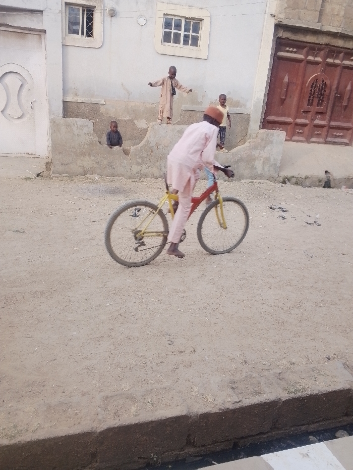 A person dressed in a light pink outfit rides a yellow and red bicycle on a dirt road. Behind them, three children stand near a short wall of a building. One child stands on the wall with arms outstretched, while the others stand or sit nearby. A large, ornate wooden door is visible to the right of the children, with windows above. A white building with simple window frames is visible in the background.