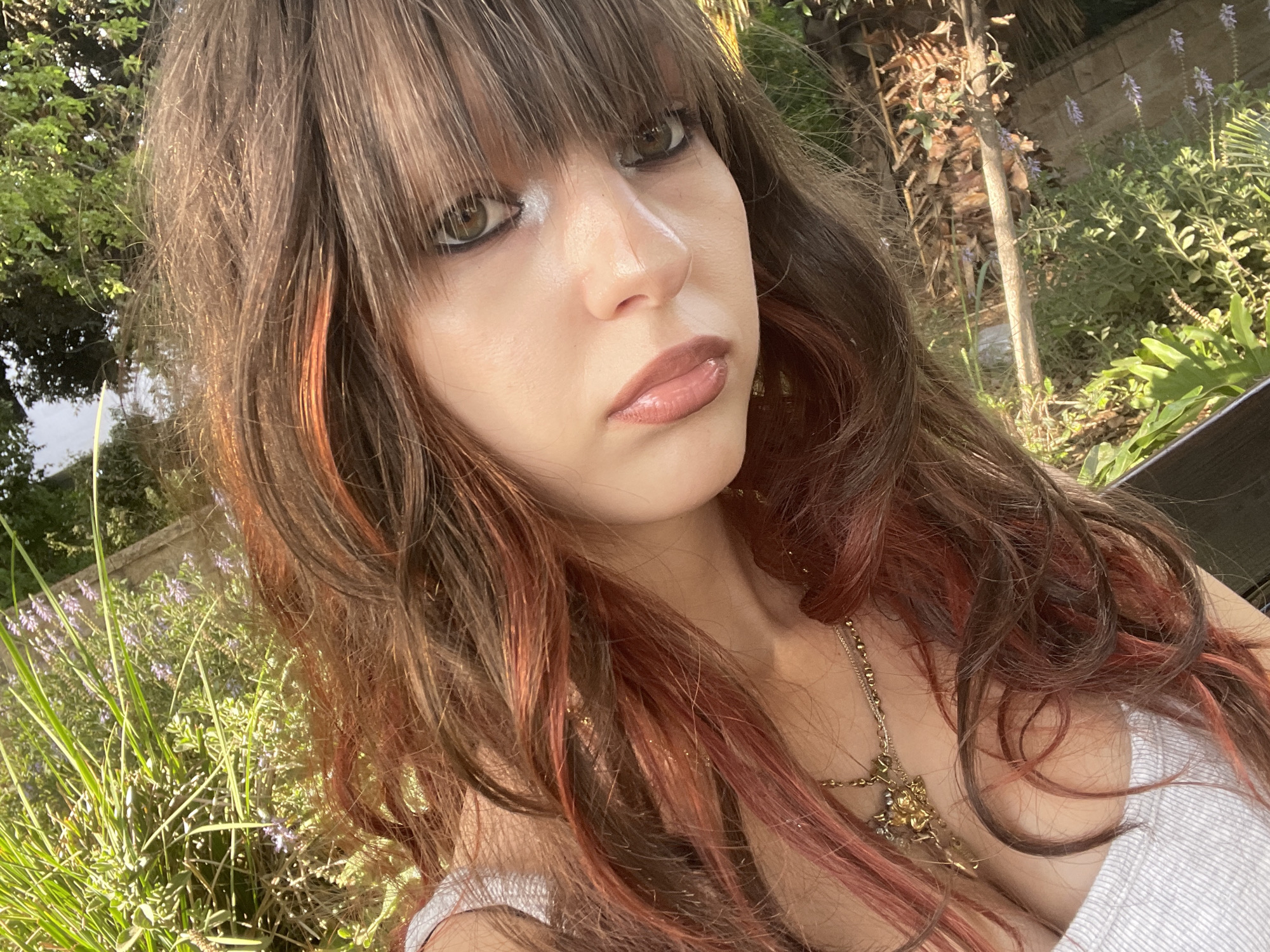 A young woman with long, wavy brown hair with reddish highlights poses for a selfie outdoors. She is wearing a white tank top and a gold necklace with a pendant. The background consists of lush green foliage and some small purple flowers, suggesting a garden or natural setting. The image appears to have been taken during daylight, with sunlight illuminating the scene. The woman's face is tilted, with her eyes looking towards the camera. Her makeup is subtle, with a focus on her lips and eyes. No specific landmarks or buildings are visible that would indicate a precise location within Raanana, Israel, though the overall environment suggests a residential or park area.