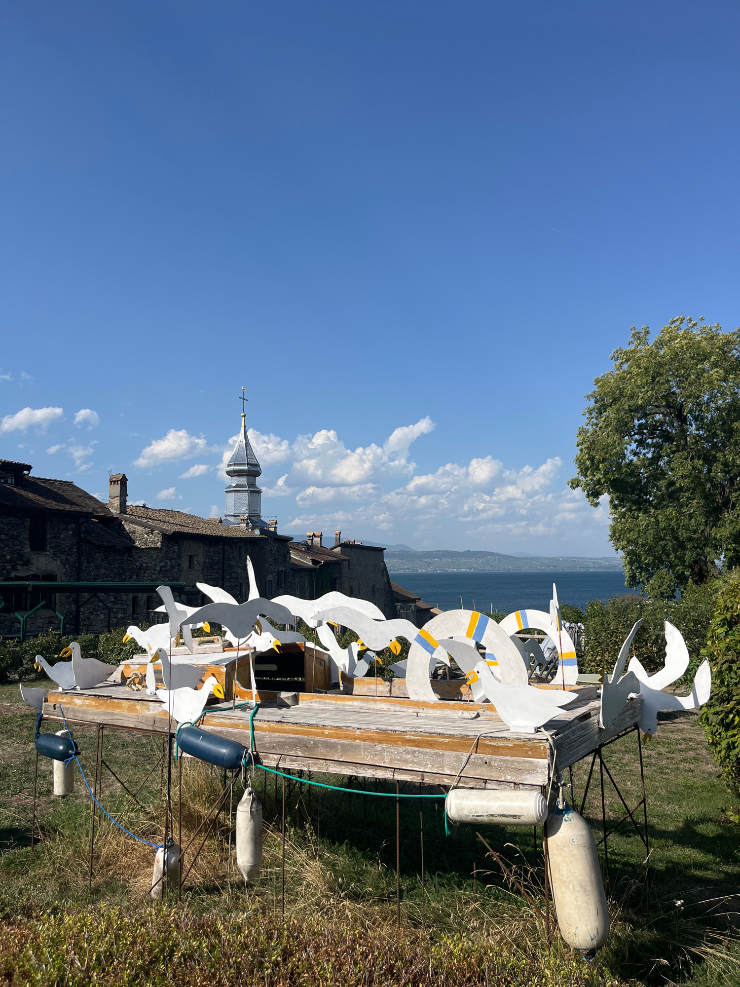 The image captures a sunny outdoor scene in Yvoire, France, featuring a whimsical sculpture of white birds and circular elements, possibly representing buoys or life preservers. The sculpture is mounted on a wooden platform supported by metal legs, with various buoys hanging from its sides. In the background, traditional stone buildings with tiled roofs and a church tower with a spire are visible. Beyond the buildings, a body of water, likely a lake, stretches into the distance, with hills and a hazy horizon line. Lush green trees and vegetation surround the sculpture and buildings, indicating a well-maintained outdoor space. The sky is a clear blue with scattered white clouds. The overall impression is one of a quaint, picturesque European village setting.