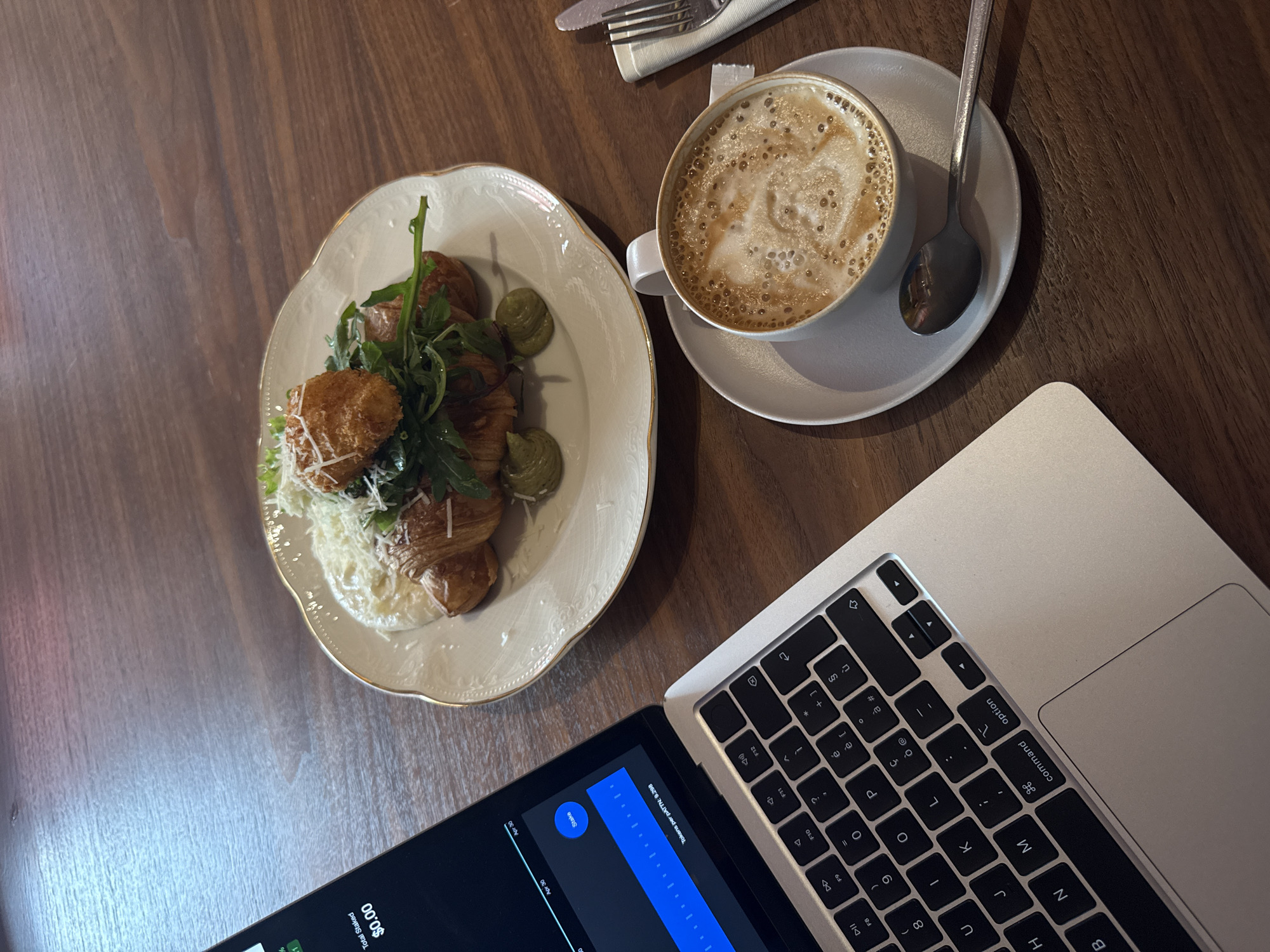 On a dark wooden table, a laptop, a plate of food, and a cup of coffee are arranged. The laptop, mostly silver in color, is open and its keyboard is visible. The plate, predominantly white with a gold rim, holds croissants with some greens and a creamy sauce. A cup of coffee rests on a light gray saucer, with a silver spoon lying beside it. A knife and fork wrapped in a cloth napkin are positioned in the upper left corner of the image. The scene appears to be set in Yerevan, Armenia.