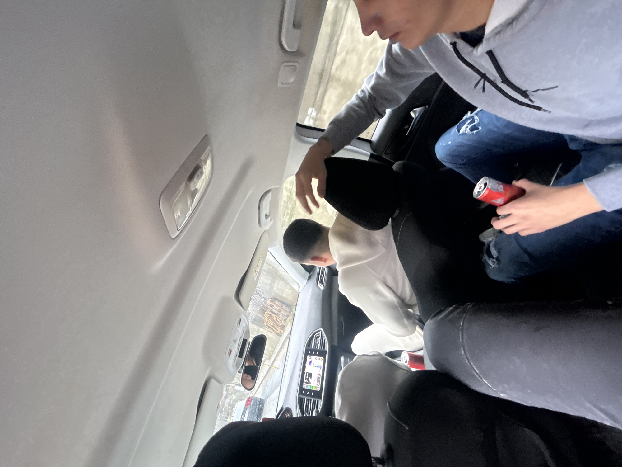 The image depicts the interior of a passenger vehicle, likely a Peugeot 308, observed from the left rear passenger's perspective, within the city of Vlahovo, Bulgaria. Four individuals are present.
In the right rear seat, a male subject wears a light blue/grey hooded sweatshirt and blue ripped jeans, holding a red beverage can. The driver, a male with short dark hair, is seated in the front left, wearing a white long-sleeved top. The front passenger, also male, occupies the front right seat, wearing a light grey long-sleeved top with his right arm extended upwards towards the ceiling. A pair of legs in dark grey pants, belonging to the fourth occupant (the photographer), is partially visible to the left of the right rear passenger's legs. A second red beverage can rests on the floor between the front and rear passenger seats on the left side.
The dashboard's infotainment screen displays a navigation map with a "533m" indicator. Through the front windshield, a grey sedan is visible ahead on a wet asphalt road, suggesting recent precipitation. Along the right exterior view, buildings with stacked wooden pallets or debris are present. The car's interior features a light grey headliner and dark seats.