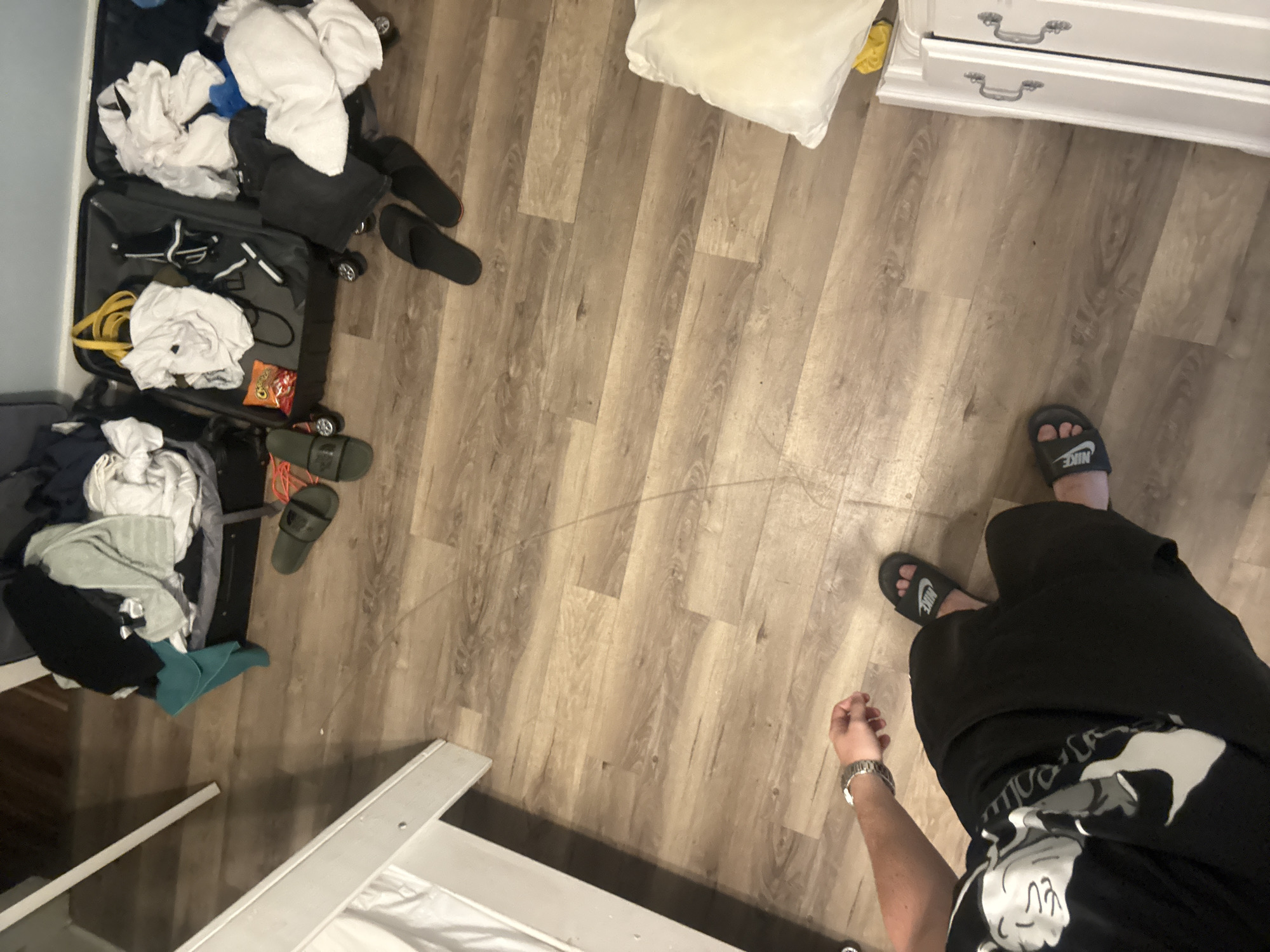 The image shows a top-down view of a room, possibly a bedroom, with light wood-grain laminate flooring. In the upper left quadrant, the bottom part of a white bunk bed frame is visible. To the right of the bed, two open suitcases are on the floor, overflowing with clothing, including white t-shirts, towels, and dark-colored garments. A yellow bag is visible inside one of the suitcases, along with a small bag of orange snacks. Several pairs of sandals are scattered on the floor around the suitcases: two dark-colored slides near the suitcases and two olive green slides further away. In the lower left quadrant, a person wearing black pants and a black t-shirt with a white graphic design is visible, along with black Nike slides. The person is wearing a silver watch on their left wrist. A large white pillow is on the floor in the lower right quadrant. To the right of the pillow, a white piece of furniture with two silver handles is partially visible. The overall scene suggests a room that is being packed or unpacked, possibly in preparation for or after a trip. The location is not determinable from the image alone.