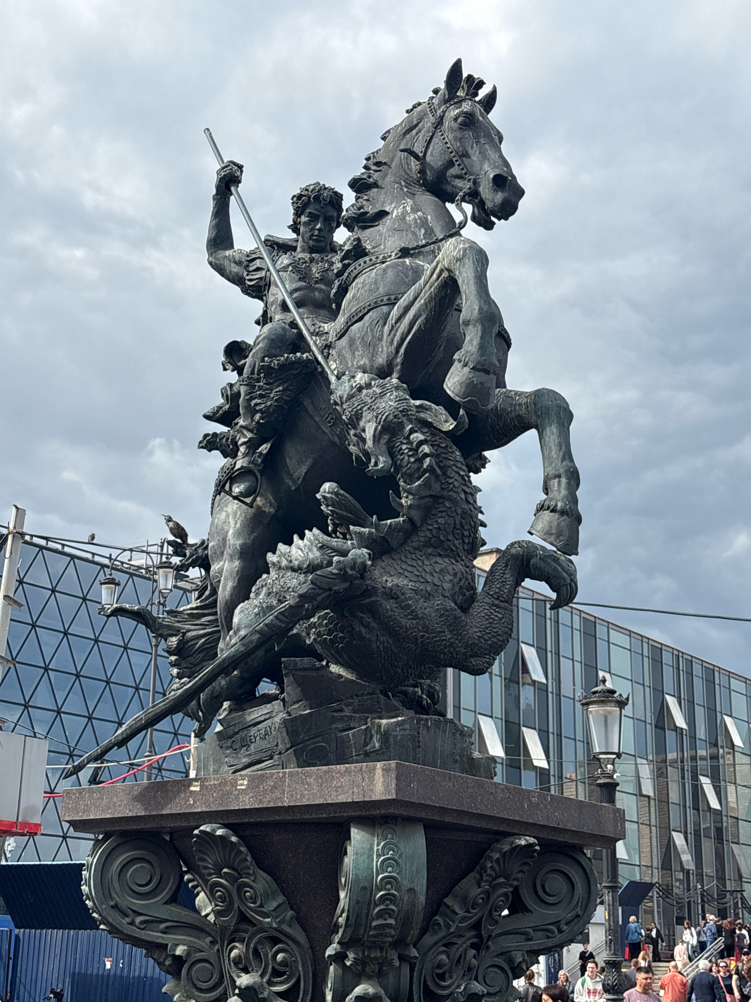This is a full-shot, eye-level, outdoor, daylight photograph of a monumental bronze equestrian statue. The statue depicts a man on horseback spearing a dragon. The man is in classical armor and has his arm raised, holding a long spear. The horse is rearing up on its hind legs, and the dragon is below it, coiled and in distress. The statue is highly detailed, with individual scales visible on the dragon's body and musculature on the horse and rider. The statue rests on a large, ornate stone pedestal.
In the background, a modern glass-fronted building with geometric patterns is visible. This building has many windows, some of which are open. A street lamp is positioned to the right of the statue, near the building. A crowd of people are visible in front of and on stairs leading up to the building. The sky above is overcast with grey clouds. The scene is the equestrian statue of Saint George, located in Moscow, Russia, in front of the State Historical Museum on Red Square.