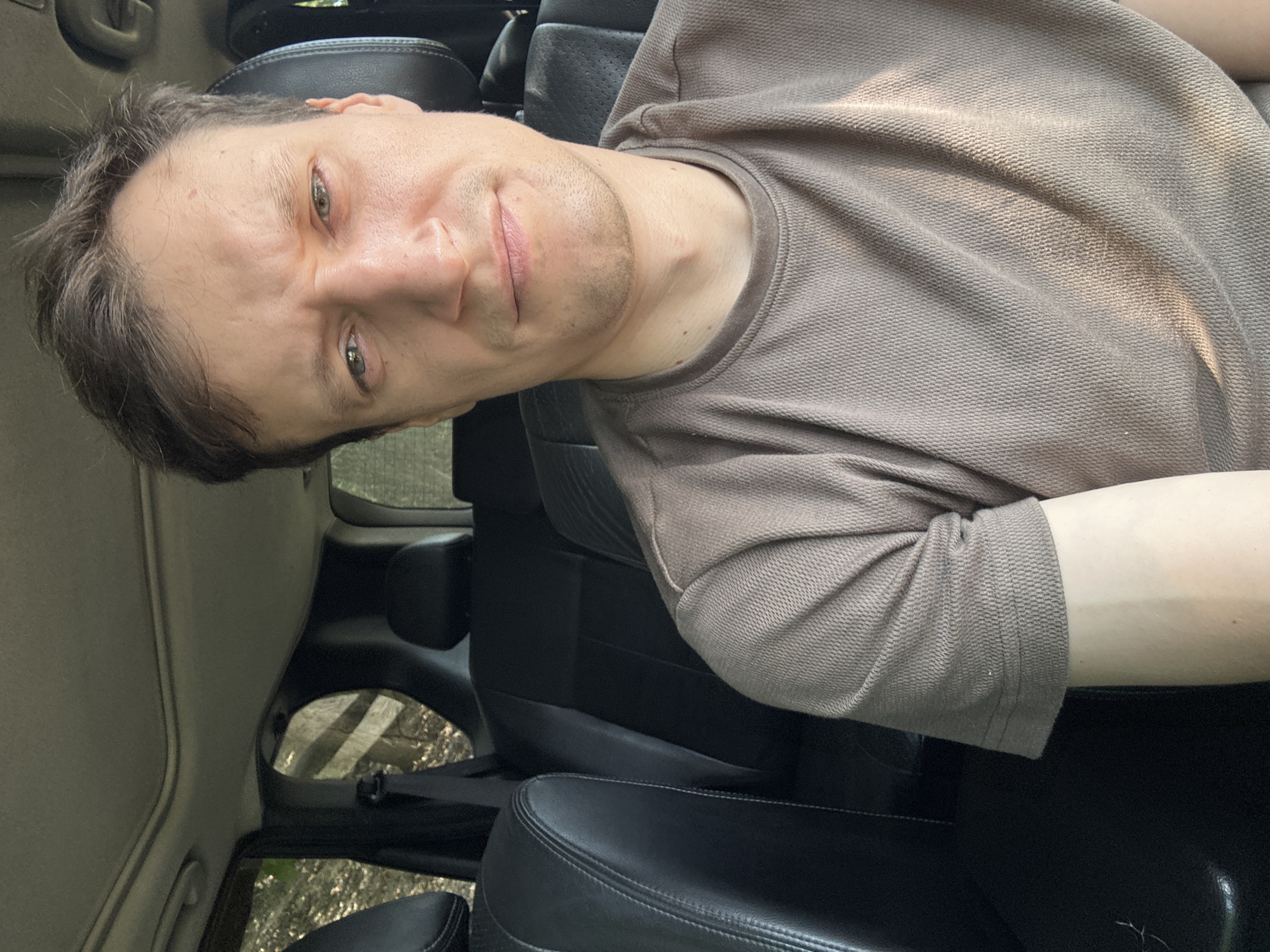 A close-up, selfie-style, medium shot captures a man sitting in the driver's seat of a car, looking at the camera with a slightly furrowed brow and a hint of a smile. He has short brown hair and is wearing a taupe-colored t-shirt with a textured pattern. The interior of the car is visible behind him, with black leather seats and a sunroof. Through the windows, trees and foliage can be seen, suggesting an outdoor setting, likely on a sunny day. The overall impression is a candid moment captured while stationary in the vehicle. The location is the city of Makiv, Ukraine.