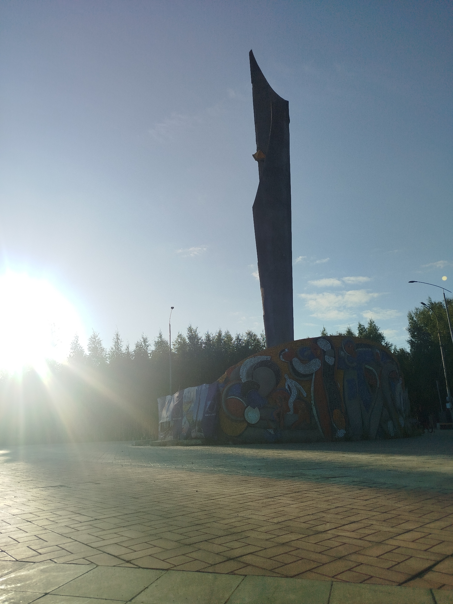 A low-angle, wide shot captures a tall, dark, abstract sculpture with a mosaic base, set against a bright, sunlit sky and a backdrop of trees in Mirny, Russia. The sun, positioned on the left side of the frame, casts strong lens flares and illuminates the scene. The sculpture itself is predominantly dark gray and has a sharp, angular form that tapers to a point at the top. The base of the sculpture is adorned with a colorful mosaic featuring abstract shapes and figures in shades of blue, orange, yellow, and white. Behind the sculpture, a dense line of evergreen trees forms a dark green wall against the sky. A street lamp with a curved arm stands to the right of the sculpture, its light source visible as a bright orb. The ground in the foreground is paved with light-colored rectangular tiles, arranged in a basket-weave pattern, reflecting the sunlight. The overall impression is one of a public monument or artwork in an outdoor setting, likely a park or square, on a clear day.