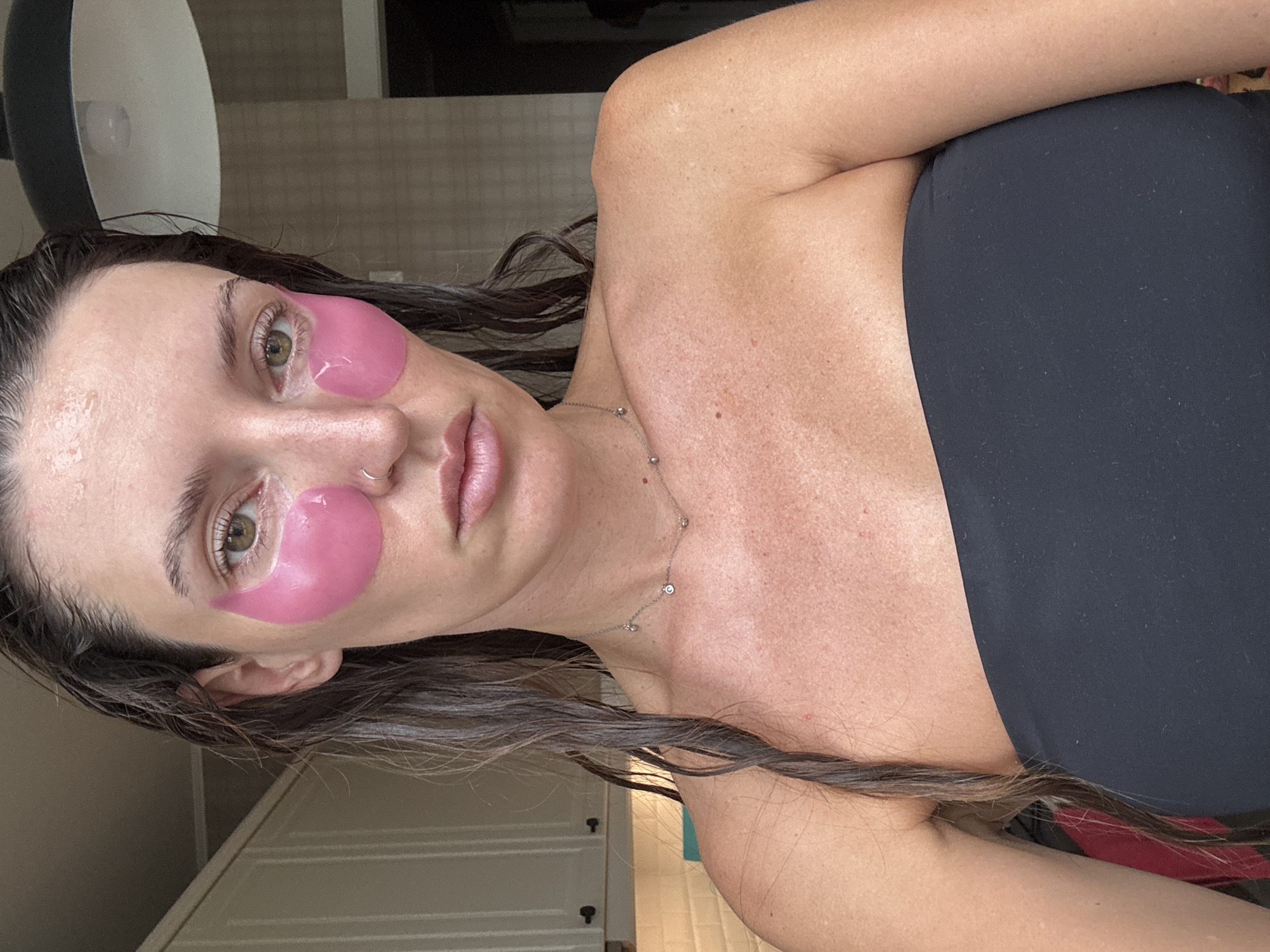 A woman with wet, dark brown hair is looking into the camera. She has pink under-eye patches and a nose ring. She is wearing a black strapless top and a delicate necklace with small diamond charms. The background shows kitchen cabinets and a tiled backsplash. This image was taken in Moscow, Russia.