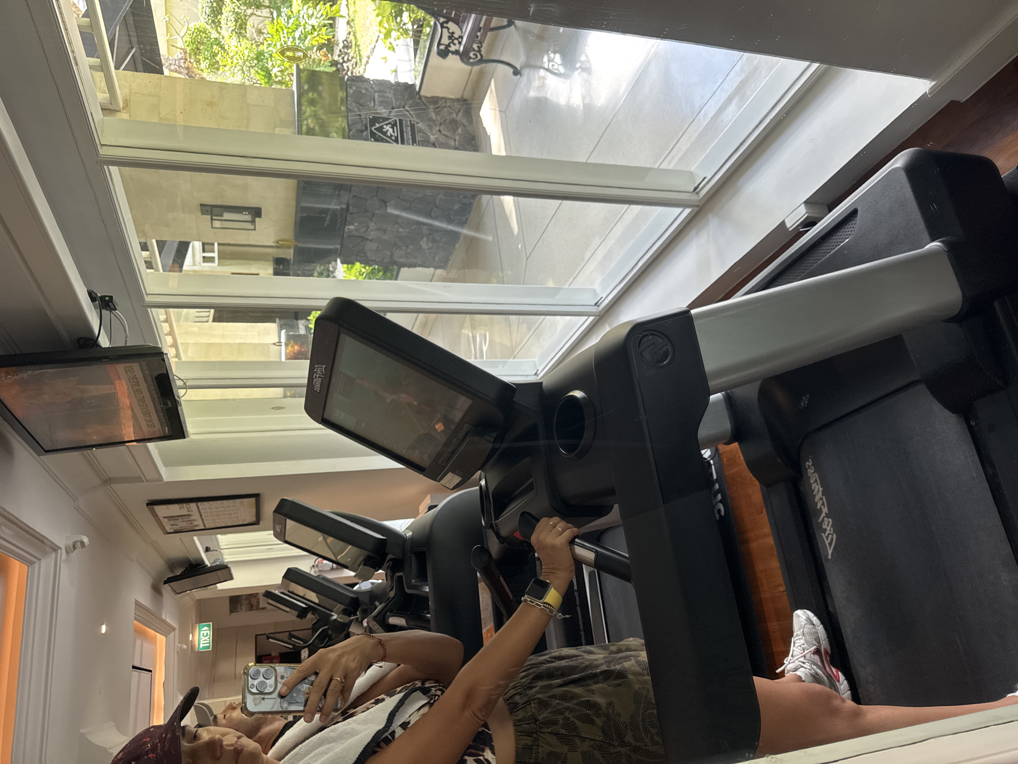A woman uses a treadmill in a gym at the Ritz-Carlton in Kuta Selatan, Indonesia. The treadmill is black with a gray handle and digital screen. The woman is wearing a camo print skirt and white sneakers. She is holding a cell phone. There are other treadmills lined up. A large window behind the treadmills looks out onto an outdoor patio.