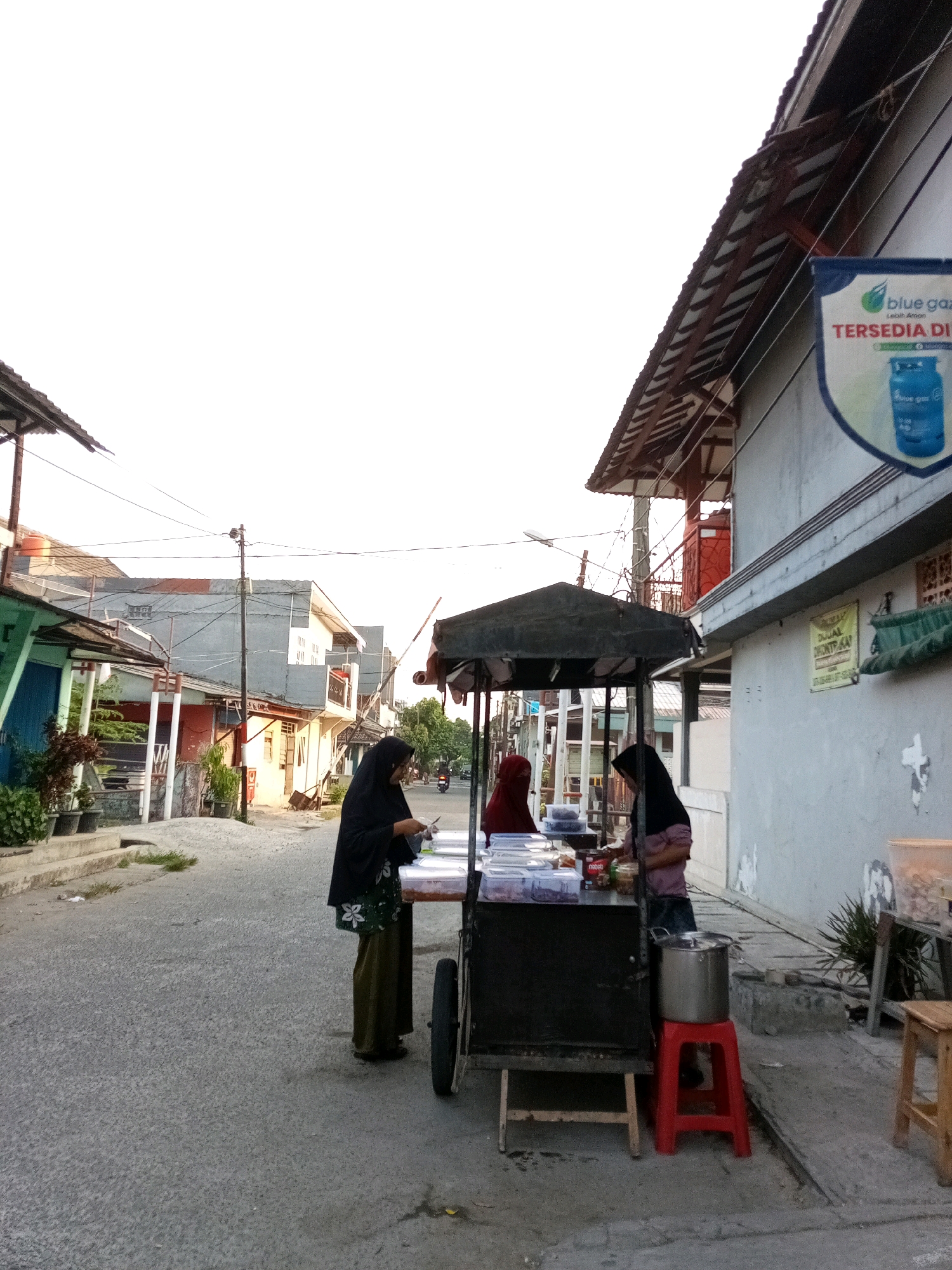 This image captures a street scene in Bekasi, Indonesia, with a focus on a food cart. Two women, dressed in Islamic attire, are interacting at the food cart, which is laden with various containers of food. One woman, wearing a black hijab and a green and white floral patterned skirt, appears to be serving or preparing food. The other woman, in a red hijab, is partially obscured but also seems to be engaged with the food items.
The food cart itself is a simple, dark-colored structure with a canopy. A large metal pot sits on a bright red plastic stool next to the cart. Various food containers, some transparent and others with colorful labels, are arranged on the cart's surface. The ground is paved with asphalt.
Behind the food cart and the women, a row of buildings lines the street. These buildings appear to be residences or small businesses, with a mix of architectural styles and colors, predominantly in muted tones. Some of the buildings have awnings and tiled roofs. A banner featuring the "blue gaz" logo and Indonesian text is visible on one of the buildings, indicating the presence of a gas retailer.
The environment suggests a residential or mixed-use area within a city. The lighting indicates it is either early morning or late afternoon, with soft, diffused light and a clear sky. The overall atmosphere is that of a typical street scene in an urban Indonesian setting, with everyday commerce taking place.