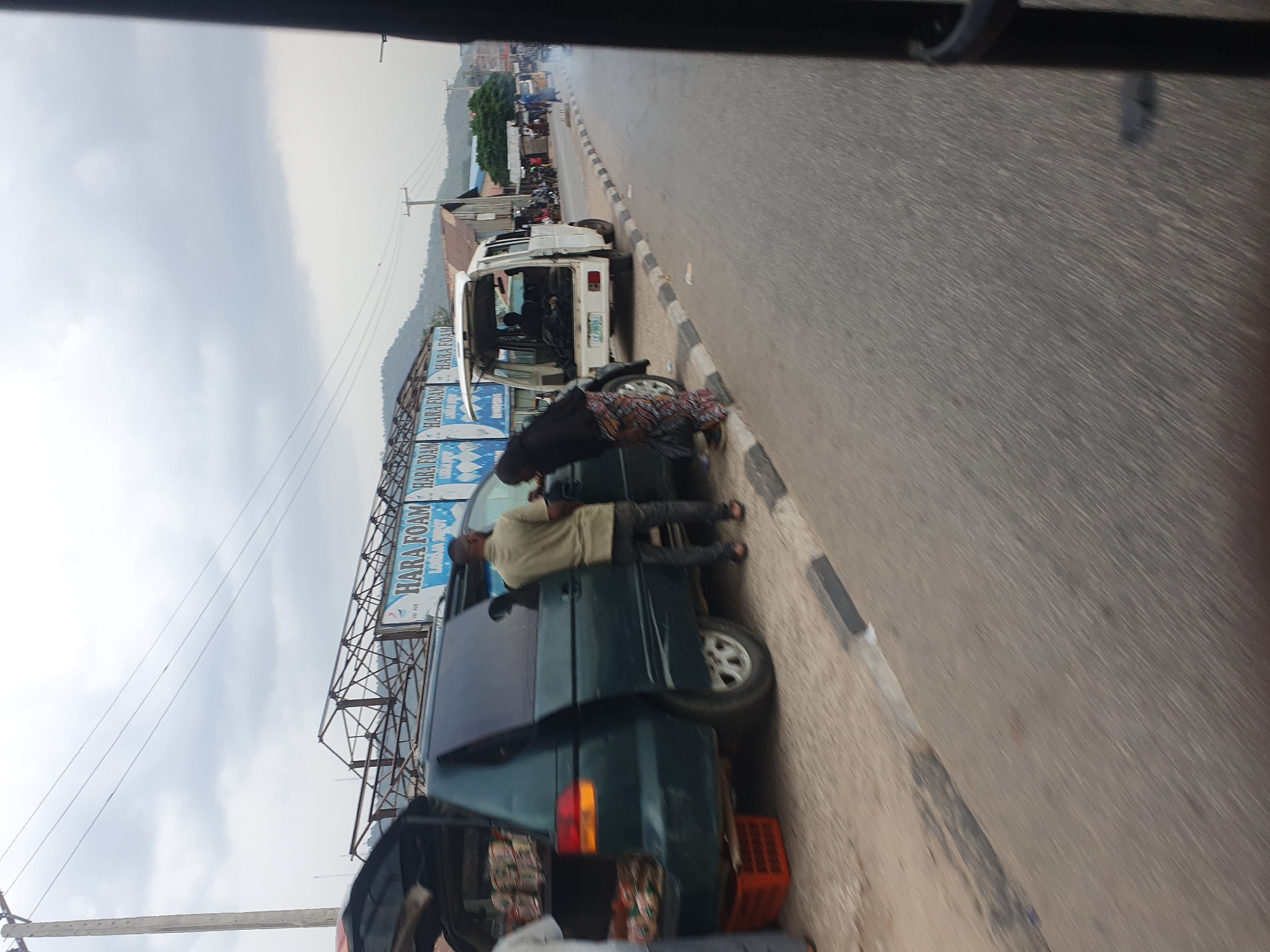 The image captures a bustling street scene in Lokoja, Nigeria, under an overcast daytime sky. The perspective is from inside a vehicle, looking out onto a roadside area.
In the foreground and midground, two minivans are parked along the side of a paved road. The closest vehicle is a dark green minivan with its rear hatch open, revealing numerous packages of goods, possibly snacks or drinks, stacked inside and in an orange crate on the ground, suggesting it's operating as a mobile stall or roadside vendor. A person in a light-colored shirt and dark pants stands next to this minivan, while another person in a patterned dress leans into its open side door. Behind this, a white minivan is also parked with its rear hatch open, further down the road, and appears to have items inside.
Along the left side of the road, a large billboard is prominently displayed, bearing the text "HARA FOAM" in large letters, with "HABAFOAM" visible below it. Beyond the vehicles and billboard, buildings line the street, and power lines extend across the frame. In the distance, a range of mountains is visible under the cloudy sky, providing a geographical backdrop to the urban or semi-urban environment. The road itself is asphalt, with a striped curb separating it from the shoulder where the vehicles are parked. Other distant, blurred figures and vehicles suggest ongoing activity further down the street. The scene depicts typical daily life and commerce in a Nigerian roadside setting.