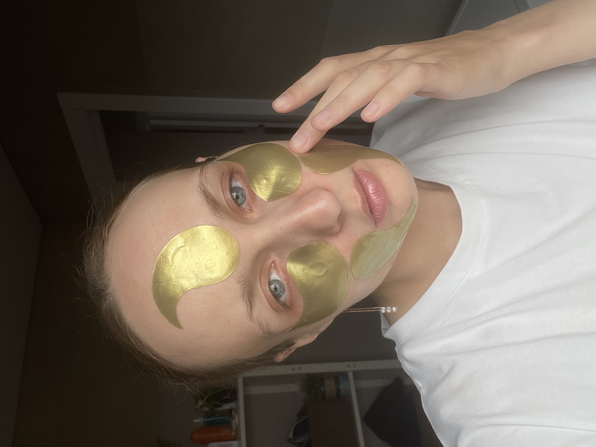 A close-up, vertically oriented shot features a person with fair skin and light blue eyes wearing gold hydrogel patches on their face, indicative of a skincare routine. The individual is looking directly at the camera with a neutral expression. Patches are visibly placed under both eyes, on the forehead above the right eyebrow, and on the left cheekbone. The person's right hand is gently touching one of the patches on the forehead.
They are wearing a simple white short-sleeved t-shirt and a delicate necklace with three small, clear gemstones around their neck. The background is minimally visible and suggests an indoor setting, possibly a room with dark walls and a light-colored shelf or furniture unit on the right side, further suggesting a home environment. No text is visible in the image. The scene conveys a moment of self-care. The location context provided is Podshivalovo, Russia.