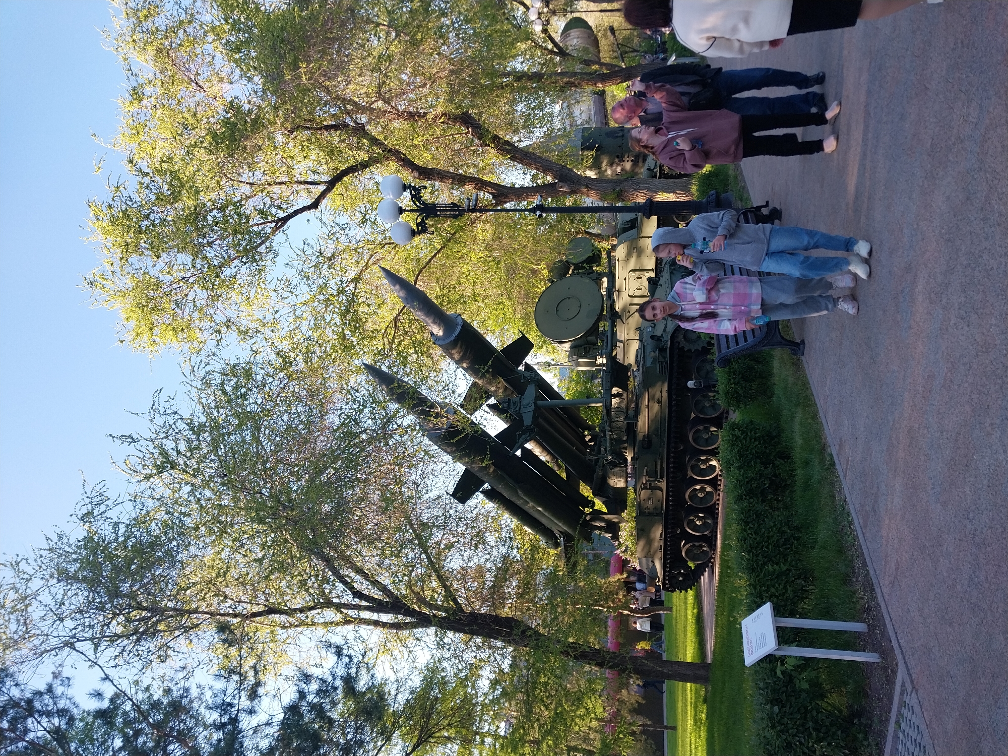 The image shows an anti-aircraft missile complex at the Victory Park in Orenburg, Russia. There are two children standing next to the missile complex. There are other people in the background. The weather is sunny, and the sky is clear. There are trees nearby. A sign is next to the exhibit.