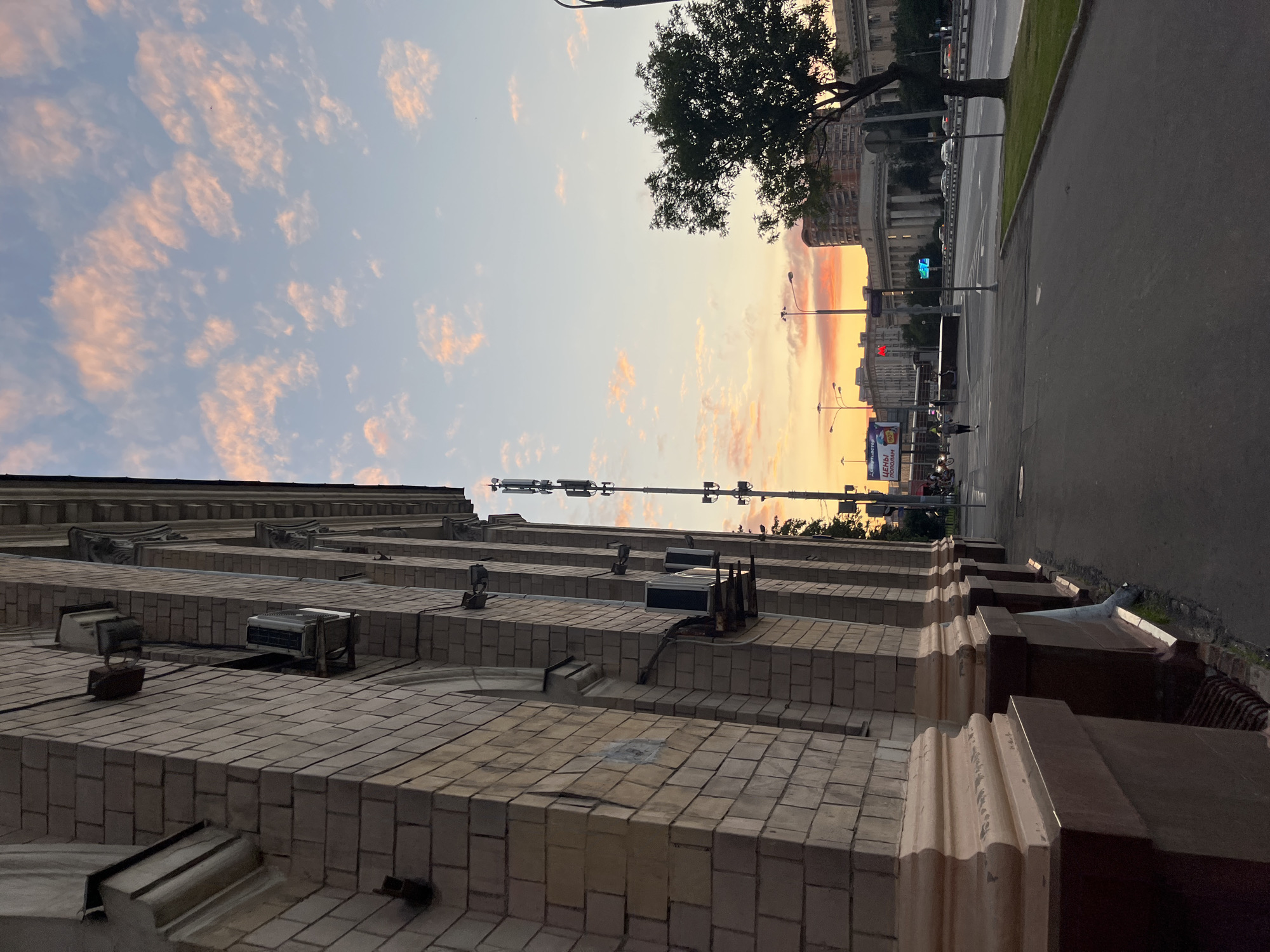 The image captures a street scene at dusk in Khimki, Russia. On the left, a brick building with arched windows and decorative columns dominates the foreground. Several air conditioning units are mounted on the building's facade. To the right, a wide street is visible with traffic, streetlights, and a dense tree. In the distance, apartment buildings and other structures line the horizon. The sky is painted with soft hues of orange and pink, indicating the setting sun, with scattered clouds. A cell tower is prominently positioned near the street. One person is walking on the sidewalk. The overall atmosphere is calm and urban, illuminated by the fading daylight and artificial lights.