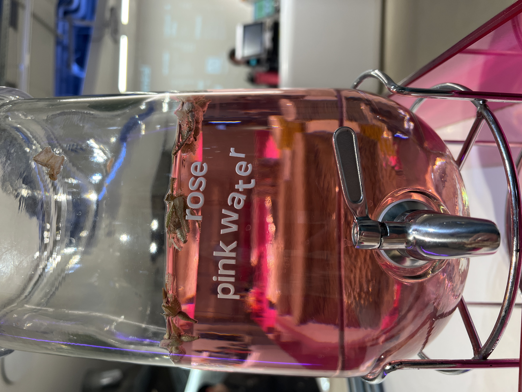 The image shows a close-up of a glass beverage dispenser containing pink liquid, labeled "rose pink water." The dispenser is housed in a metal frame and features a chrome tap. There are botanical inclusions along the interior edge of the glass where the label is located. The location may be Moscow, Russia; however, there is no evidence to support this claim.