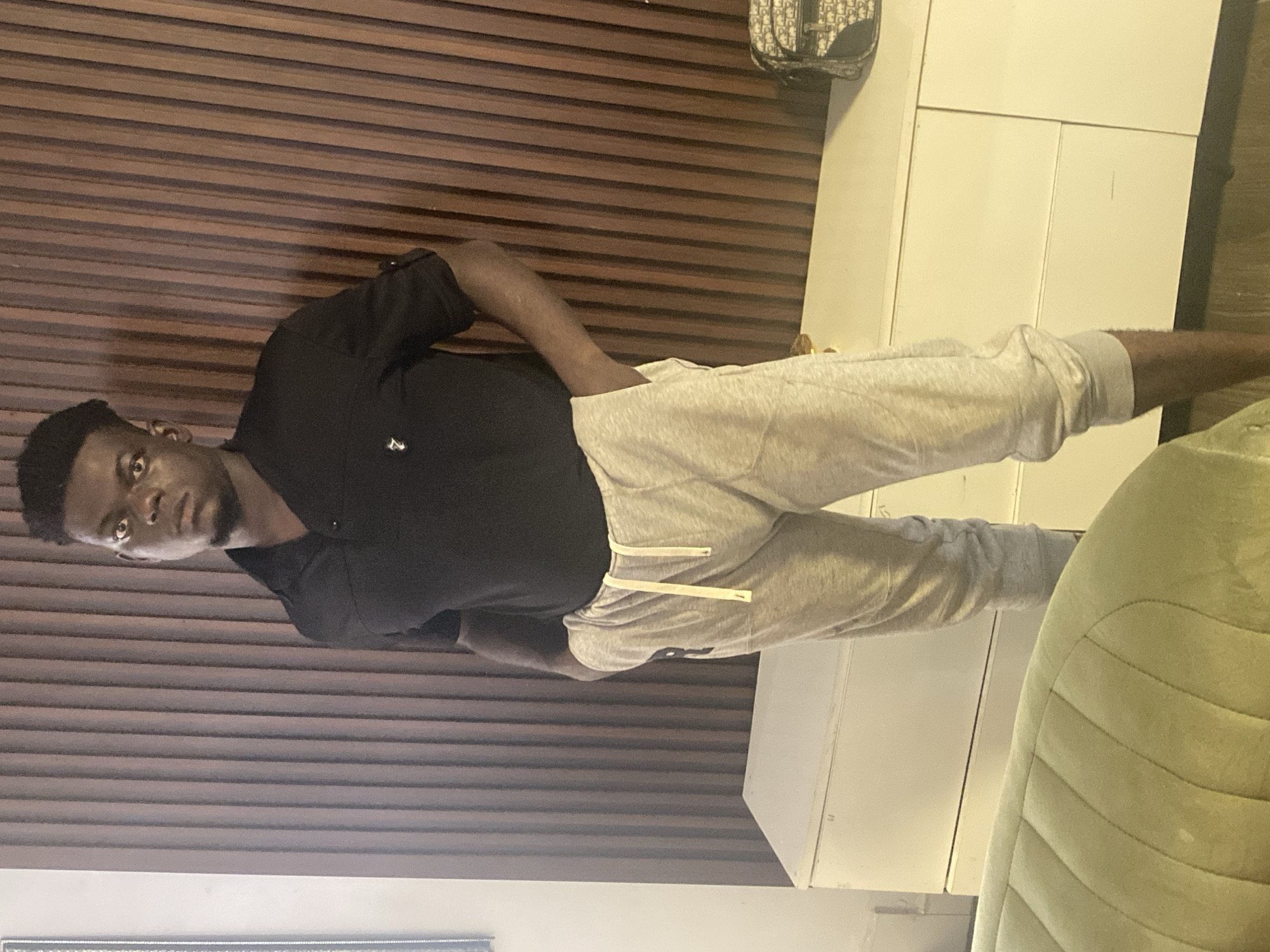 A young Black man is standing indoors, looking directly at the camera. He is wearing a black short-sleeved polo shirt and light grey sweatpants with a white drawstring. His right hand is in his pocket. The background features a wall covered in horizontal brown wooden slats or panels. To his right, there is a white, multi-paneled cabinet or storage unit. In the upper right corner, a white and black patterned bag, possibly from the brand Dior, is visible. Part of a light green, cushioned chair or couch is seen in the bottom right corner of the frame. The overall setting suggests a modern indoor space, potentially a living area or lounge. The man's expression is neutral. The lighting appears to be artificial, and the time of day is not discernible.