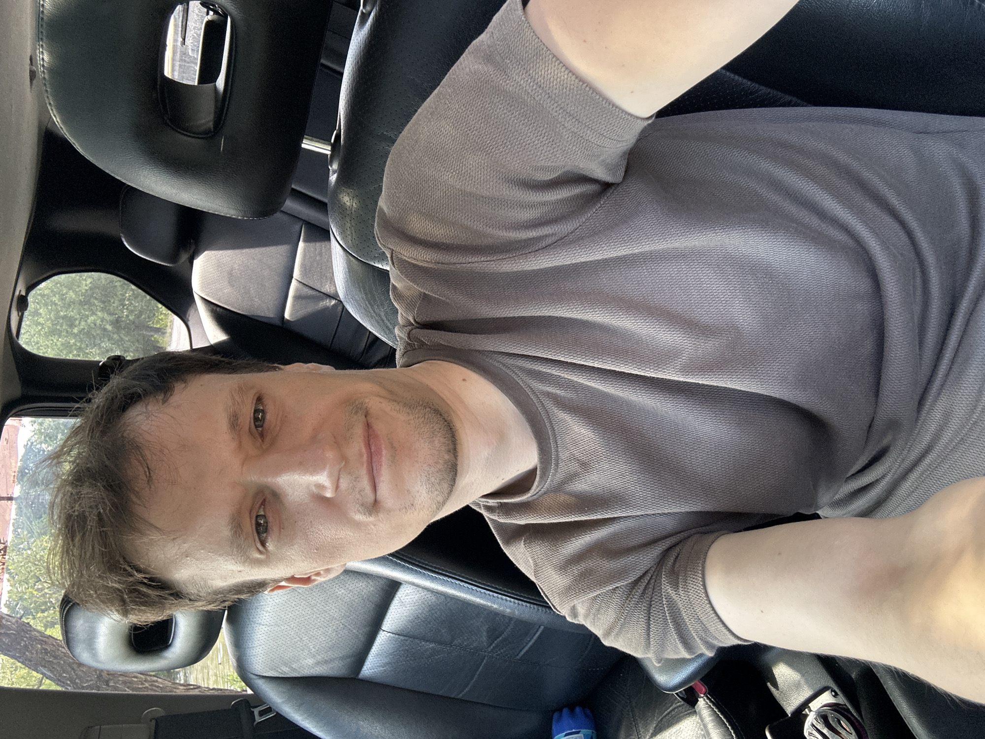 A selfie of a man in the driver's seat of a car, facing the camera with a slight smile. He has light brown hair, light skin, and is wearing a grey t-shirt. The car's interior, with black leather seats and headrests, is visible. Through the windows, trees and foliage can be seen, suggesting an outdoor location. The scene is lit by natural sunlight. The image does not contain any information about the location in Makiv, Ukraine.
