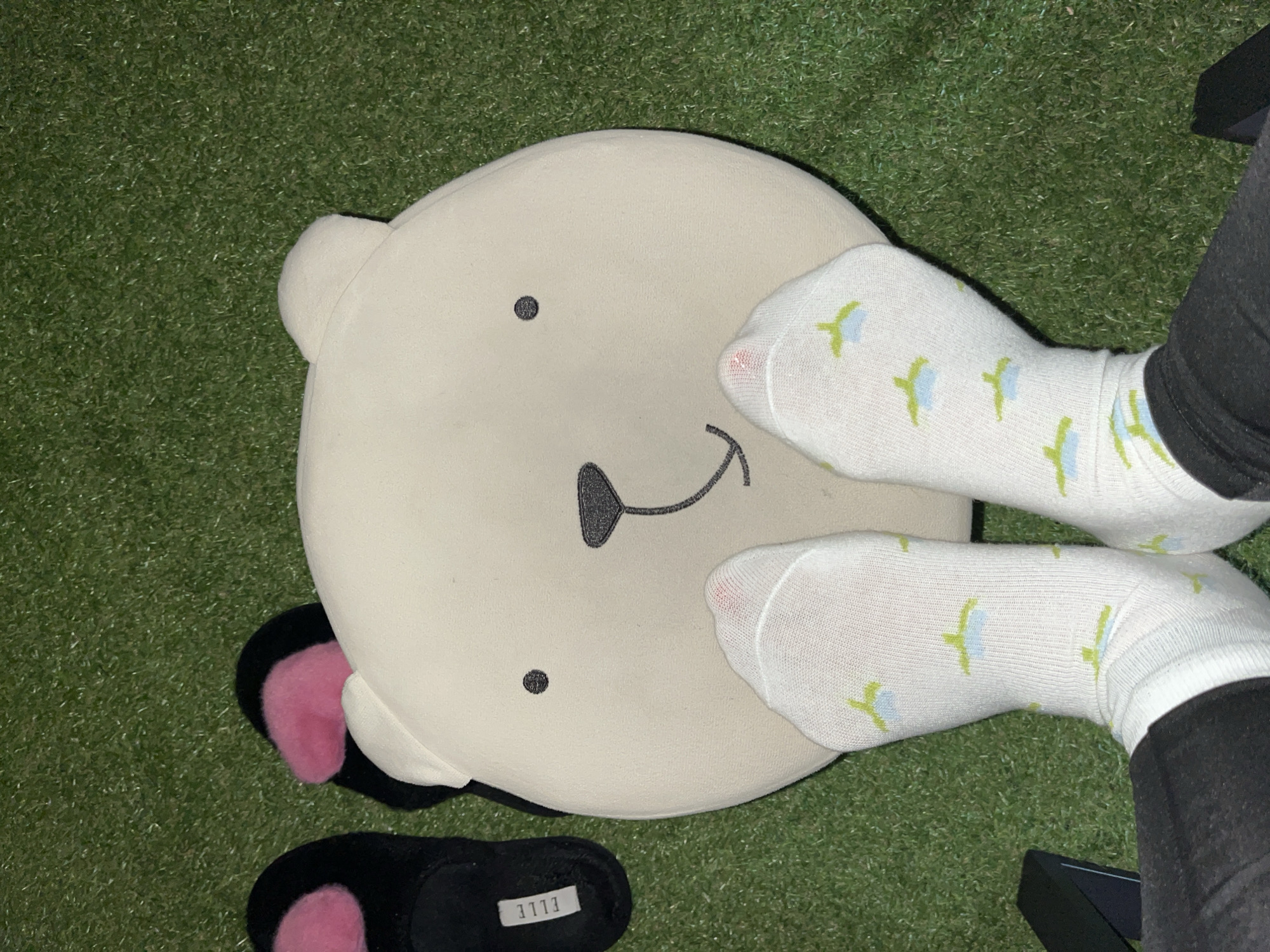 The image depicts a close-up, overhead view of a person's feet wearing white socks with a green and blue pattern, resting on an artificial grass surface. To the left of the feet are black slippers with pink accents. In the center, a large, beige, bear-shaped pillow is positioned, partially obscuring the feet. The pillow has simple embroidered features, including two black dots for eyes and a stitched mouth. The scene is indoors, as indicated by the lighting and the presence of indoor slippers and a pillow. The artificial grass suggests an indoor setting designed to mimic an outdoor environment, possibly in a home or recreational area. There is no indication of Garmisch-Partenkirchen, Germany, within the visual information presented.