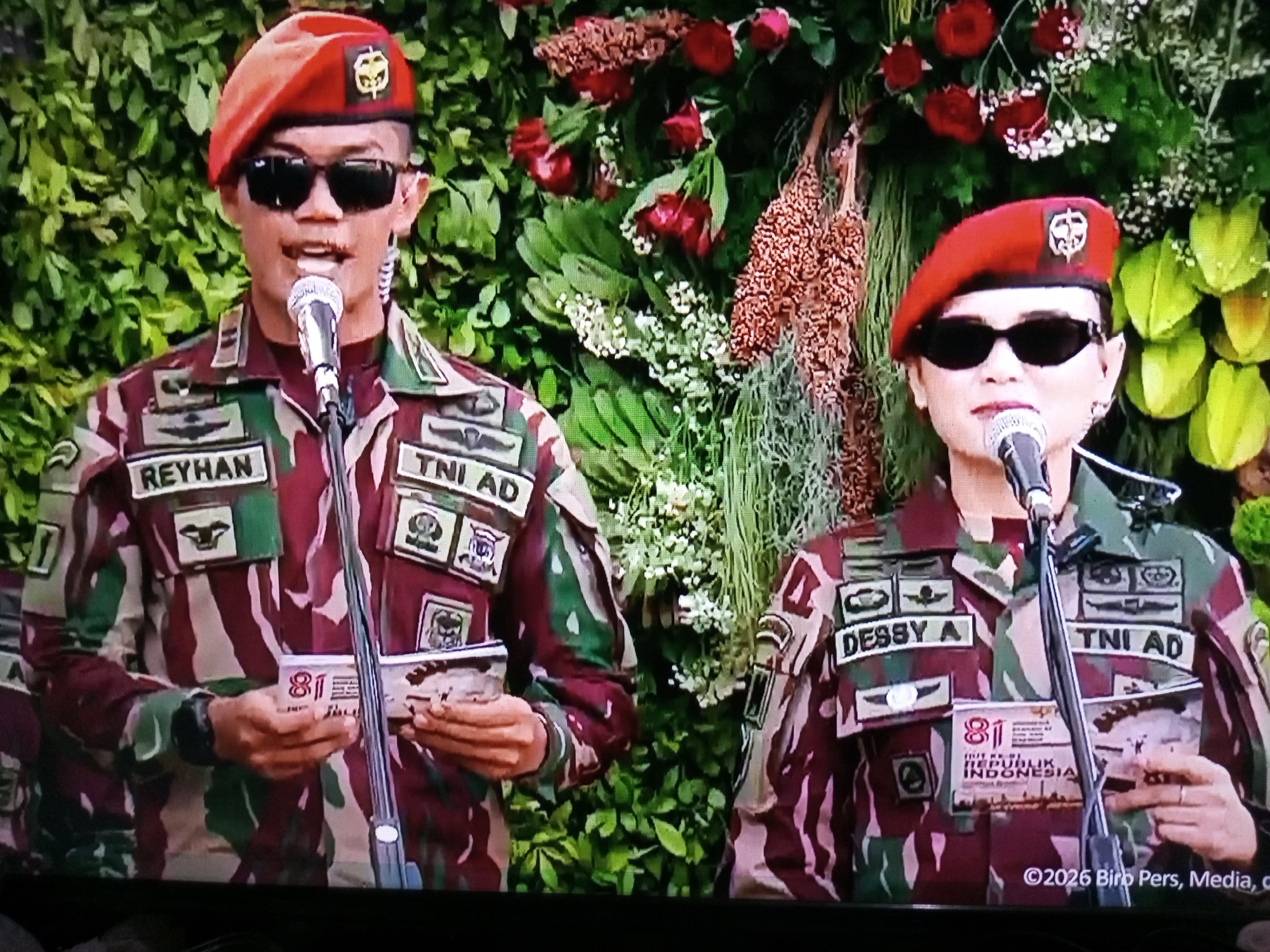 Two individuals in military uniforms are standing in front of a floral backdrop, likely at an official event in Banyudono, Indonesia. The individual on the left, identified by the name tag "REYHAN," wears a red beret and sunglasses. They are speaking into a microphone. The individual on the right, identified by the name tag "DESSY A," also wears a red beret and sunglasses, and is holding a document with the number "81" visible. Both are wearing camouflage uniforms with various patches, including "TNI AD" on their sleeves. The backdrop consists of lush green foliage, red roses, and other floral arrangements. A watermark at the bottom right of the image reads "©2026 Biro Pers, Media, O."