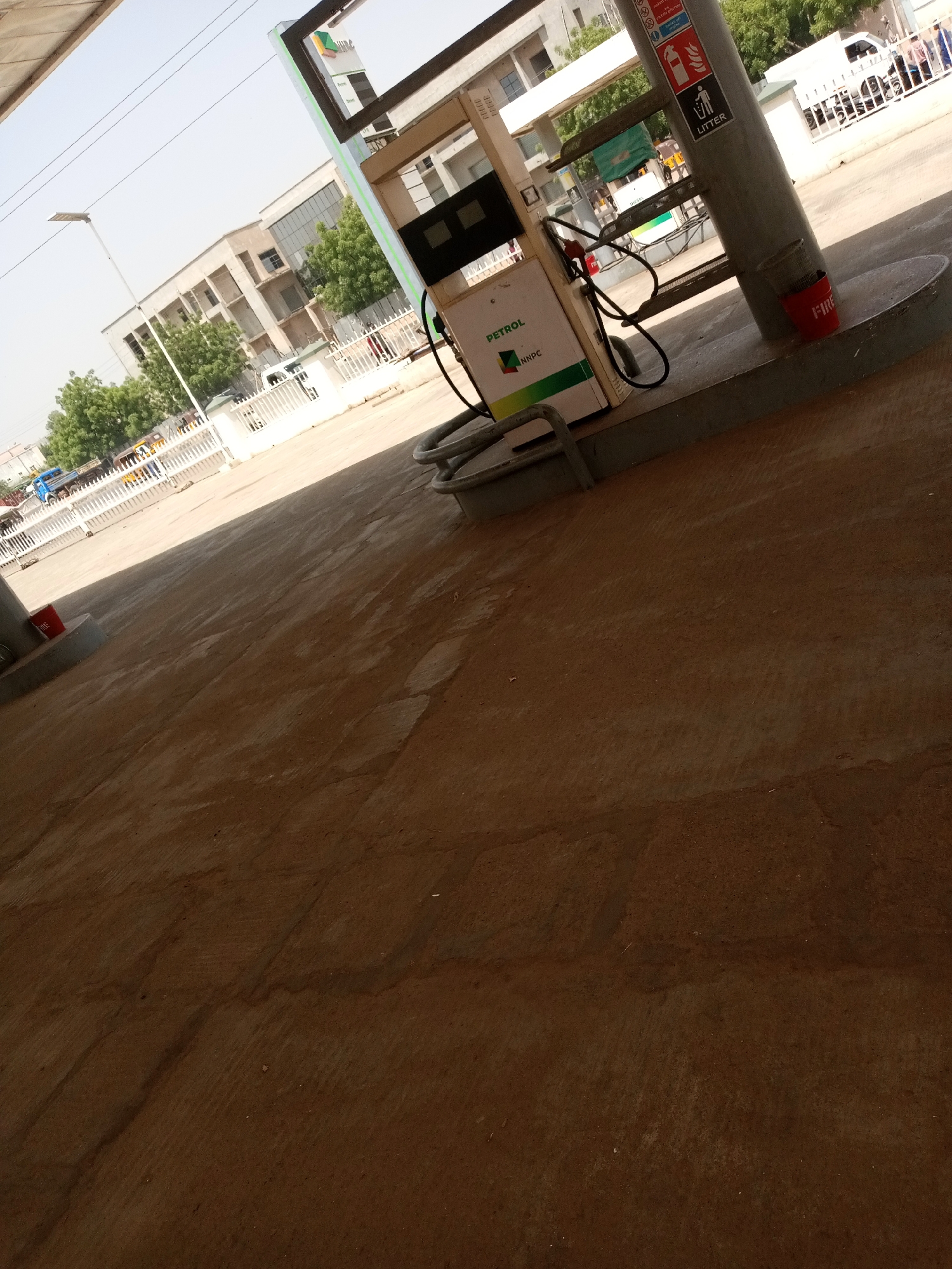The image shows a gas station with a petrol pump. The pump has the "NNPC" logo and the word "PETROL" visible on it. A red fire bucket is placed near the pump. In the background, there is a white fence, trees, and buildings. A white van and other vehicles are also visible behind the fence. There are also signs on the pump post, one with icons of a fire extinguisher, a warning symbol, and a litter bin. The scene appears to be outdoors during the day, with bright lighting suggesting sunny weather. The ground is paved with concrete. The angle of the photo is from a low perspective, looking up towards the gas station and the surrounding area.