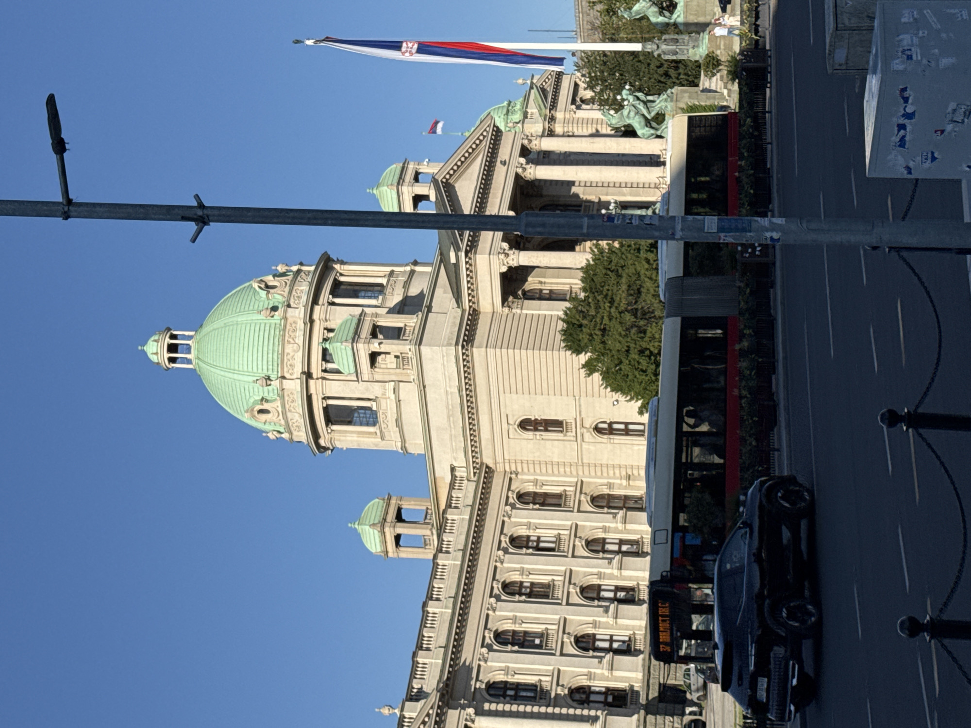 This is a photograph of the National Assembly of Serbia building in Belgrade, Serbia. The building is a large, neoclassical structure with multiple domes and columns. In front of the building, a bus is driving down a street, and a black SUV is parked on the side of the road. A lamppost is in the foreground, and a Serbian flag is flying on a flagpole. The sky is clear and blue.