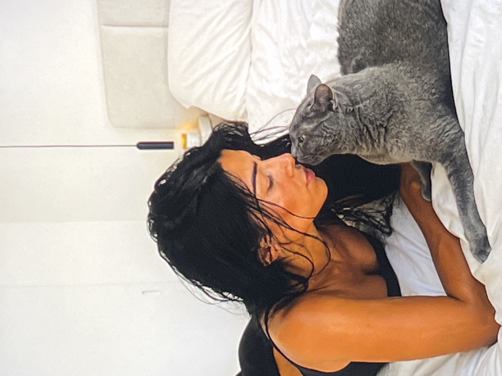 An overhead medium shot captures a woman with long, dark hair lying on a white bed and nuzzling a gray cat. The woman is wearing a black tank top and is on her side, facing the cat. The cat is also on its side, facing the woman, and their noses are touching. The bed is covered with a white comforter, and a white headboard is visible in the background. A bedside table with a lamp and a small jar is to the right of the woman. A modern black pendant light hangs from the ceiling above the bed. The overall lighting is bright and airy, suggesting a bedroom setting. The scene appears to be indoors, within a residential environment.
