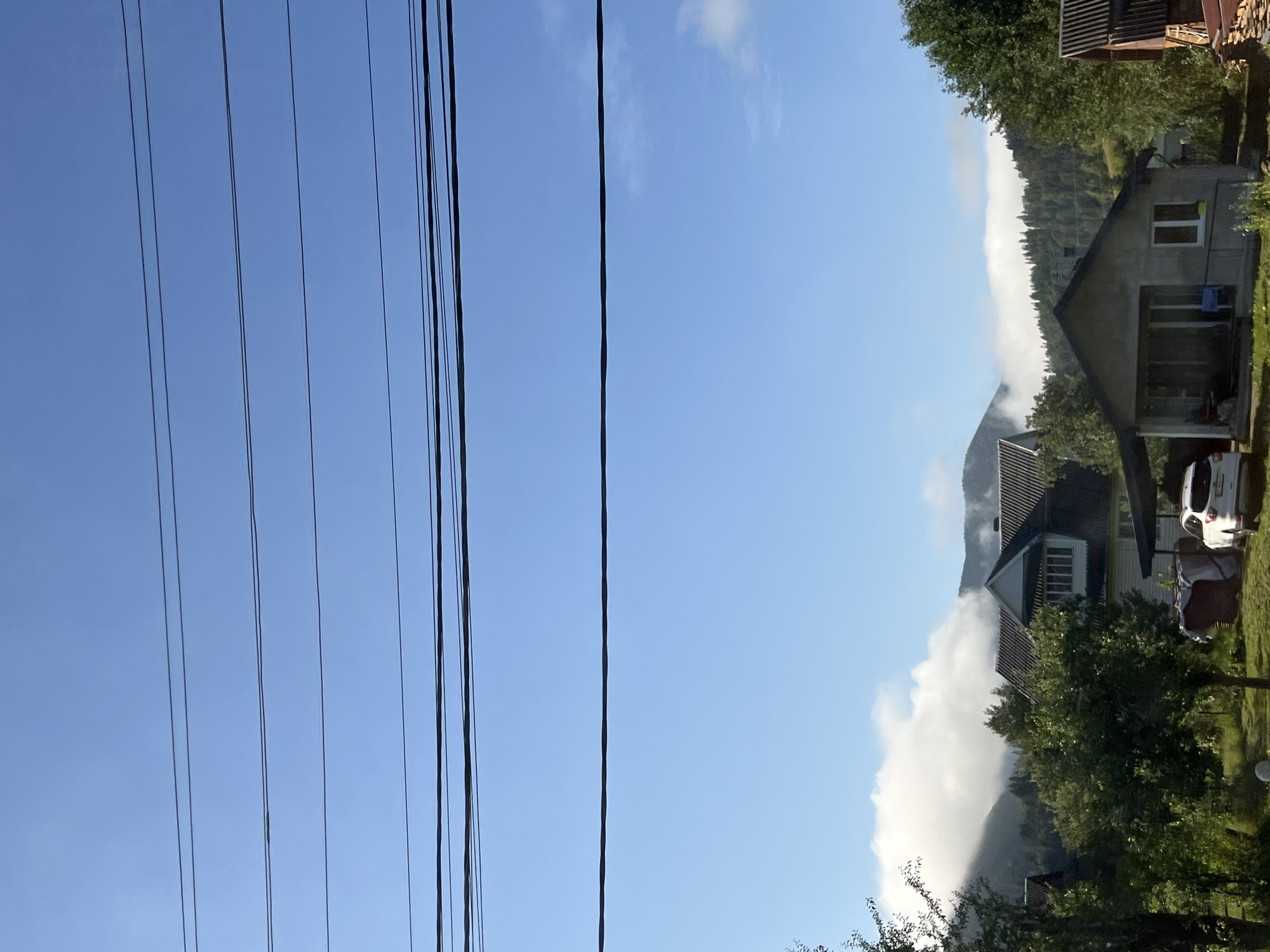The image shows a rural landscape with several houses and electrical wires against a bright blue sky. In the foreground, there are trees and a white car parked under a carport. A white house with a dark roof is visible behind the car. Further in the background, a larger house with white siding and a dark roof is prominent, partially obscured by trees. Mountains are visible in the distance, with clouds rolling over them. The overall scene suggests a tranquil, possibly mountainous, residential area. The location is identified as Tatariv, Ukraine.