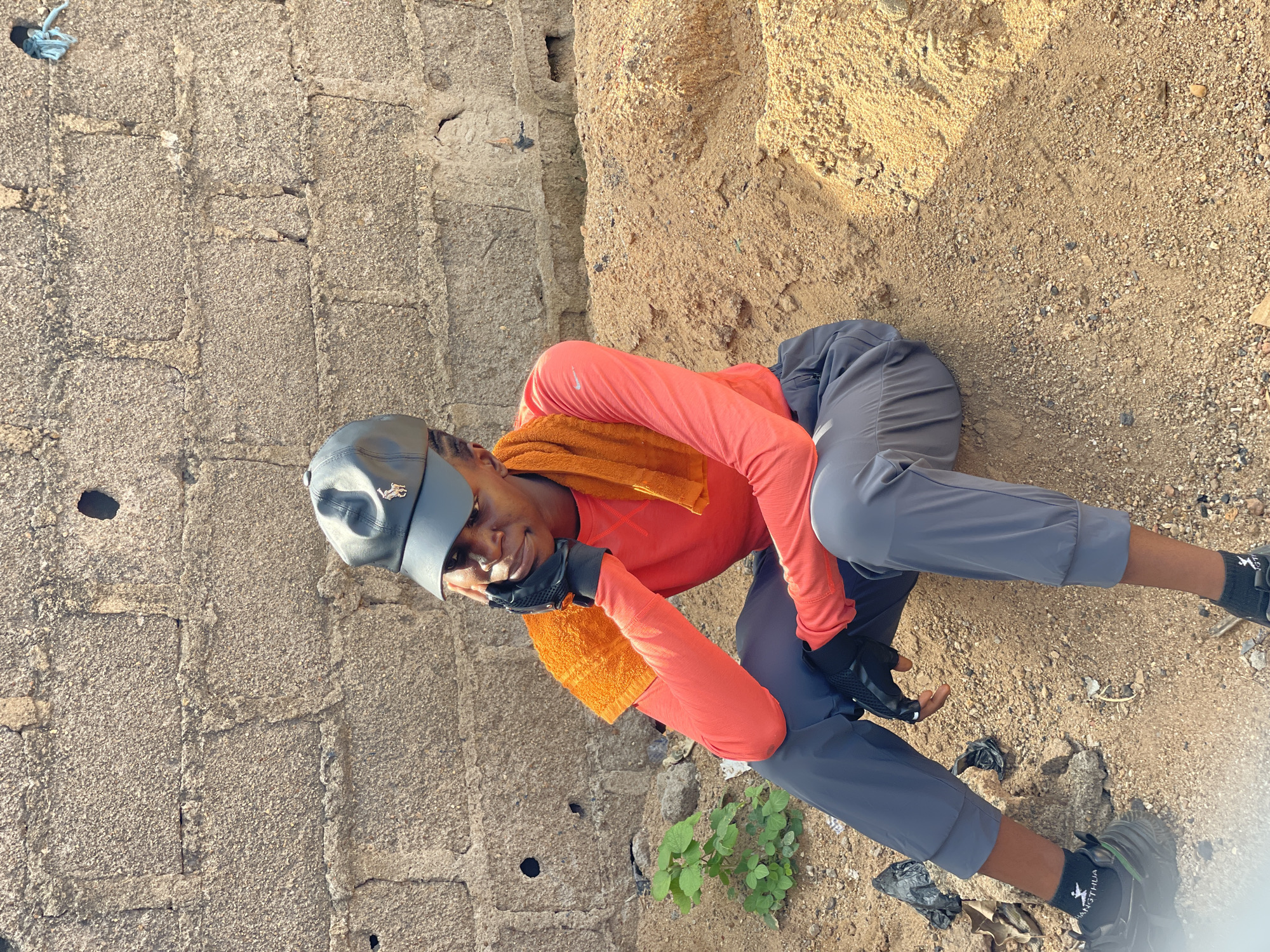 A young man is sitting on the ground in Akwanga, Nigeria. He wears a gray baseball cap, an orange long-sleeved shirt, gray athletic pants, black gloves, and black shoes with white lettering on black socks. An orange towel is draped around his neck. He is smiling, his face visible, and he appears to be resting his head on his hands. To his left is a brick wall, while to his right, the ground is sandy and rocky. A small green plant grows near him.