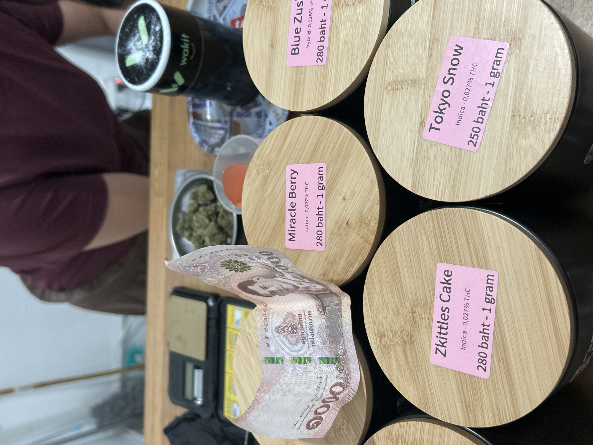 The image shows a close-up view of a counter in what appears to be a cannabis dispensary in Ratsada, Thailand. On the counter, there are several black cylindrical containers with wooden lids, each labeled with a pink sticker indicating the strain of cannabis, its THC content, and price. Some of the strains visible are "Miracle Berry," "Blue Zushi," "Zkittles Cake," and "Tokyo Snow." A Thai 1000 baht banknote is draped over one of the containers. To the left, a digital scale is visible, suggesting the products are being weighed. In the background, out of focus, a person wearing a burgundy shirt is partially visible, along with some shelving and what looks like a cleaning mop. The overall scene suggests a retail environment where cannabis products are sold.