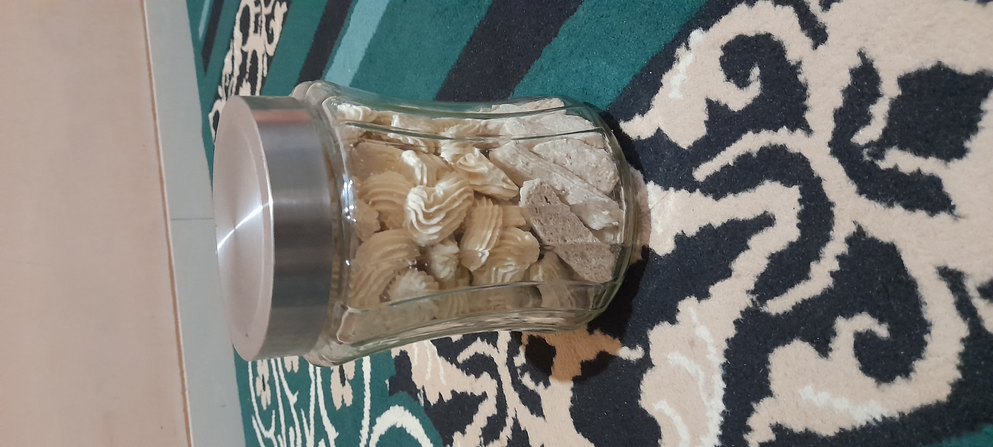 The image shows a clear glass jar filled with cookies, placed on a carpet with a black and cream patterned design. The jar has a silver lid. The cookies inside are of two types: some are piped into swirled shapes, and others are cut into irregular rectangular pieces. The background consists of a plain wall and a portion of a green and black striped carpet. The lighting appears to be from an overhead source, casting a shadow from the jar onto the carpet. The scene is an indoor setting, likely a domestic environment, with no indication of a specific location within Dawe, Indonesia, beyond it being a generic home interior. There are no people or actions depicted in the image.
