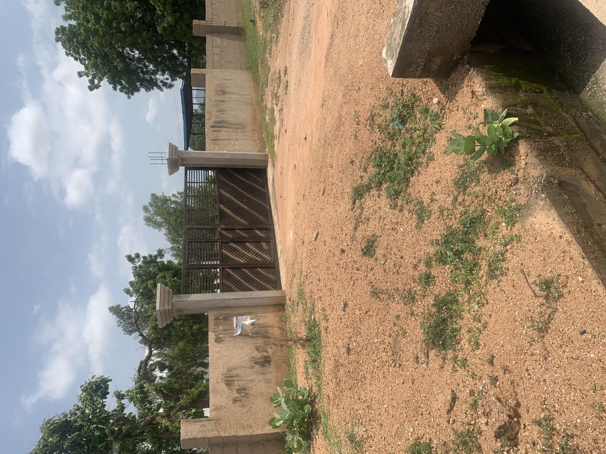 The image depicts a dirt road leading to a closed metal gate. The gate is set within a textured, light brown wall, with two stone pillars flanking the gate. To the left of the gate, there is a patch of green plants. The wall extends to the right, constructed of concrete blocks in the distance. Behind the wall, trees and a covered structure are visible. The ground is covered with orange sand and sparse green vegetation. The sky is blue with scattered white clouds. The scene appears to be located in Jalingo, Nigeria, possibly in a residential or industrial area.