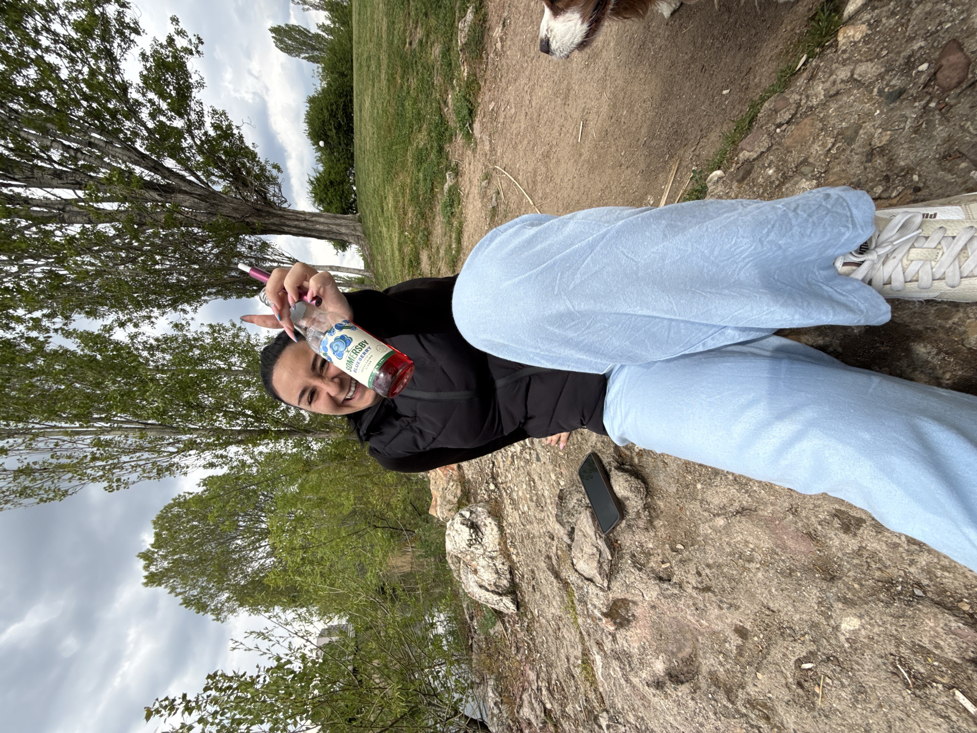 The image, rotated 90 degrees clockwise, depicts a person relaxing outdoors in what appears to be a park or natural area in Sofia, Bulgaria. A female-presenting individual is seated on a rocky, dirt-covered surface, smiling brightly and looking towards the camera. She wears a black puffer jacket and light blue, loose-fitting pants, with white sneakers visible on her right foot, which bears a partially discernible "Puma" logo.
In her left hand, she holds a clear glass bottle containing a reddish beverage, clearly labeled "SOMERSBY BLUEBERRY." Alongside the bottle, she also holds a slender, pink and purple vape pen. Her fingers are painted with light pink nail polish, and she forms a "peace sign" gesture. Her dark hair is pulled back from her face.
The background features large trees with abundant green foliage, indicative of a verdant outdoor setting. The sky is overcast or partly cloudy, suggesting daytime. In the upper right corner, the head and neck of a brown and white dog are visible. A dark-colored smartphone rests on the rocky ground next to the person. The scene conveys a relaxed and happy mood in a natural environment.