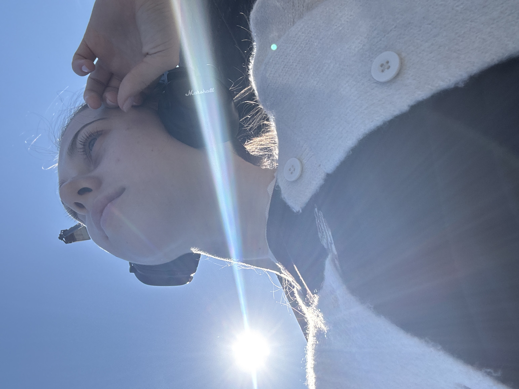 A low-angle, outdoor, eye-level shot captures a person from the chest up, looking towards the sun. The person has fair skin and is wearing a white cardigan with two visible buttons and a black undergarment. They are also wearing black headphones, one of which has the "Marshall" logo. Their right hand is raised to their ear, touching the headphone. Sunlight glints brightly from the upper left of the frame, creating lens flare and highlighting the person's hair and face. The background is a clear blue sky. The image does not contain any information that would indicate a specific location within Miltenberg, Germany.