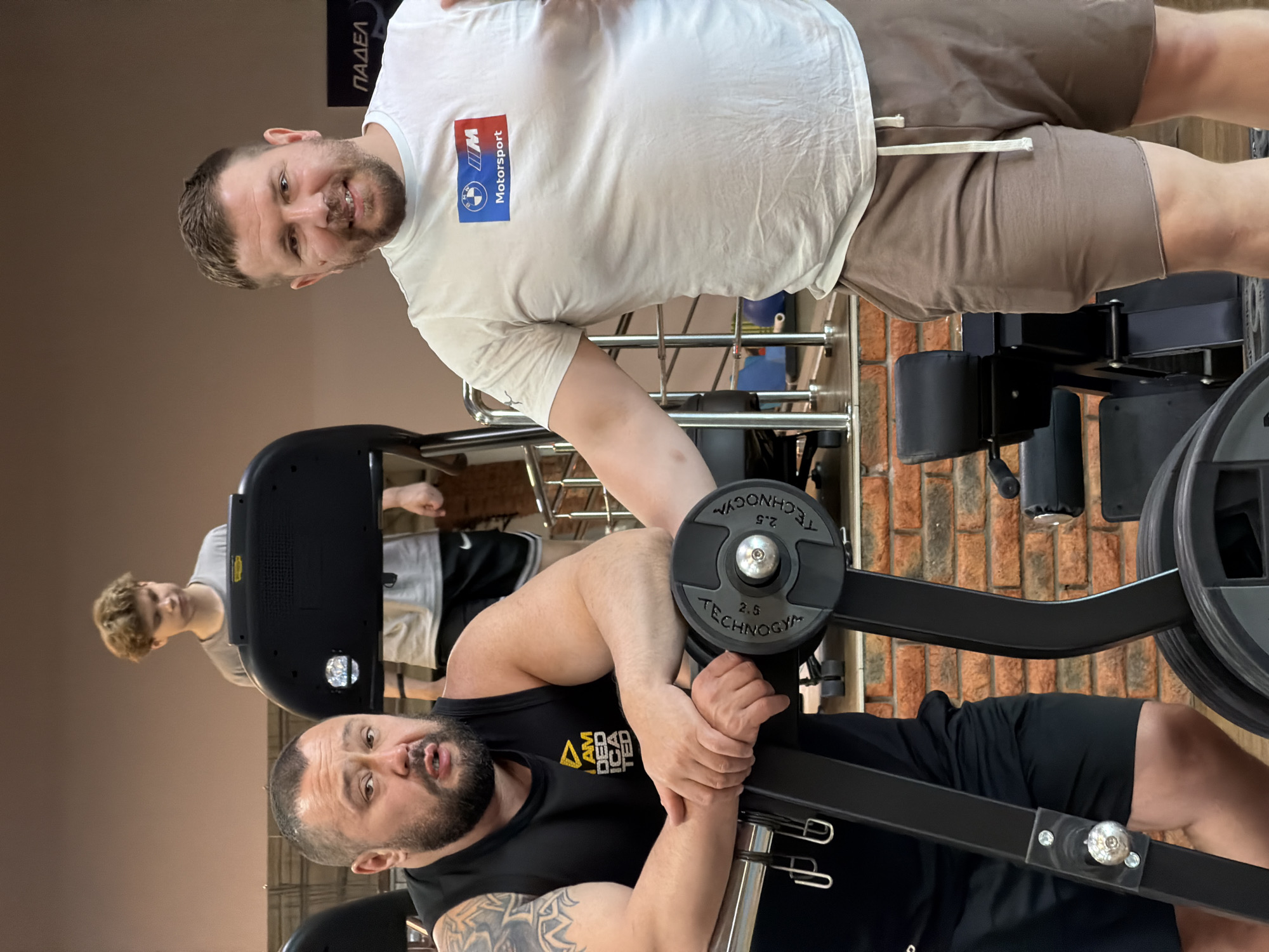 The image shows two men in what appears to be a gym in Chaikovsky, Russia. The man in the foreground is lying on a bench press machine, lifting weights. He is wearing a black tank top with a triangle logo and has a beard and visible tattoos on his arm. The other man, standing beside the bench, is wearing a white t-shirt with BMW Motorsport logo and brown shorts. He is looking at the camera with a smile. In the background, a third person is visible on a treadmill. The gym has brick walls in the background.