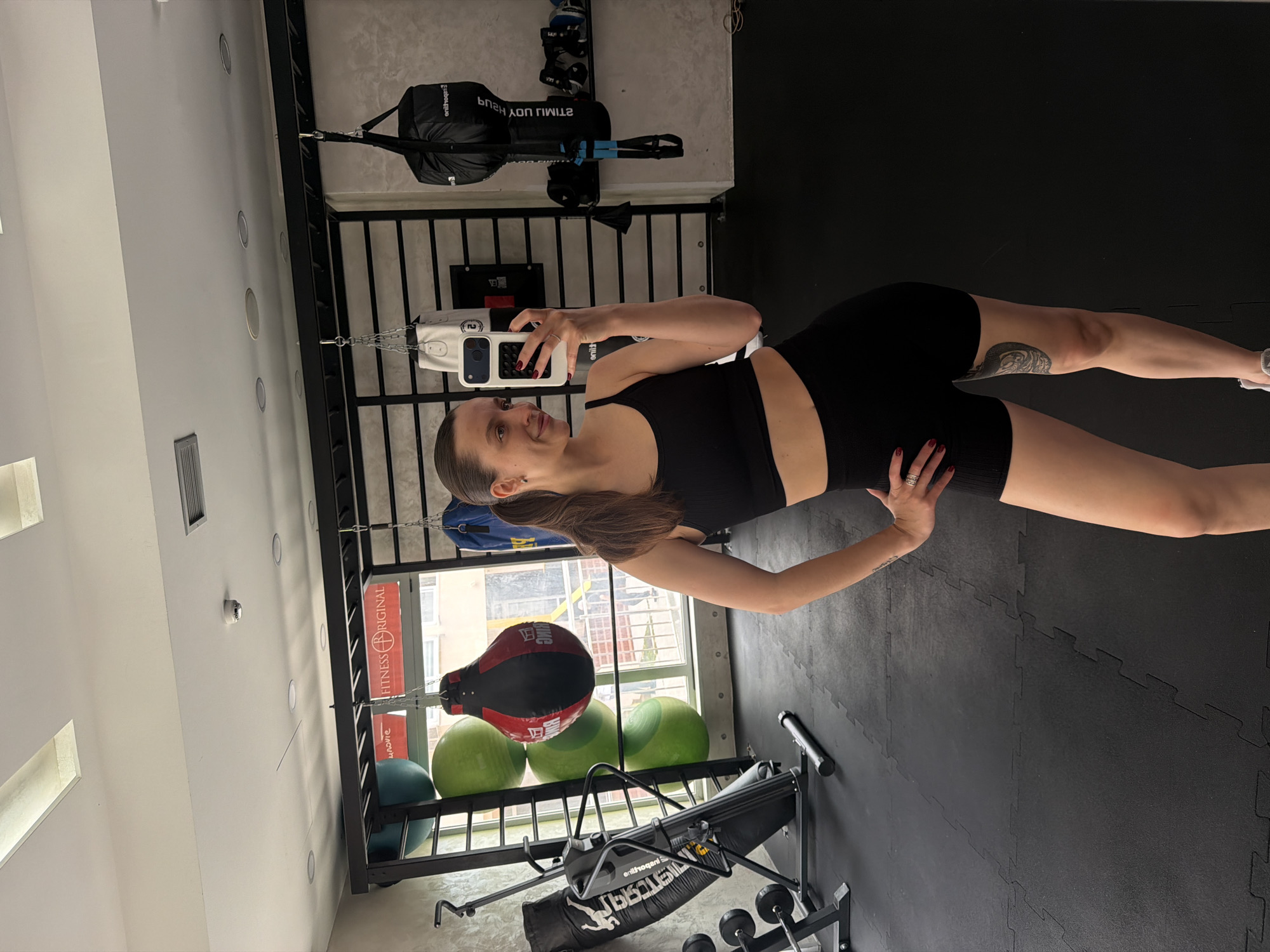 A woman is taking a selfie in a gym. She is wearing a black sports bra and black shorts. The gym has a black and gray floor, a white wall, and a black rack with various exercise equipment. In the background is a window showing a view of Budva, Montenegro.