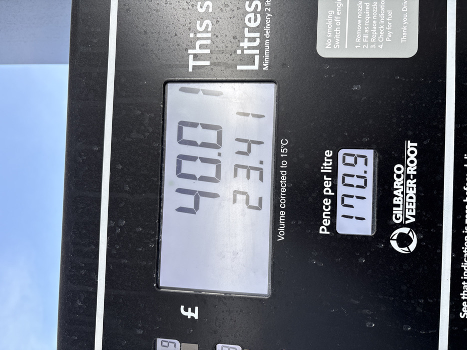 A close-up image depicts the digital display screen of a Gilbarco Veeder-Root fuel pump, located at a service station in Bayston Hill, United Kingdom, under a clear blue sky. The dark grey pump surface shows minor surface dust or water spots. The primary display indicates a total cost of £40.01 and a dispensed volume of 23.41 litres, with the text "Volume corrected to 15°C" below the litre count. A separate display shows the fuel price at 170.9 pence per litre. To the right, a white panel provides safety instructions: "No smoking Switch off engine," followed by a four-step guide for fueling: "1. Remove nozzle 2. Fill as required 3. Replace nozzle 4. Check indication Pay for fuel." The panel concludes with "Thank you. Driv". No individuals or vehicles are visible in the frame.
