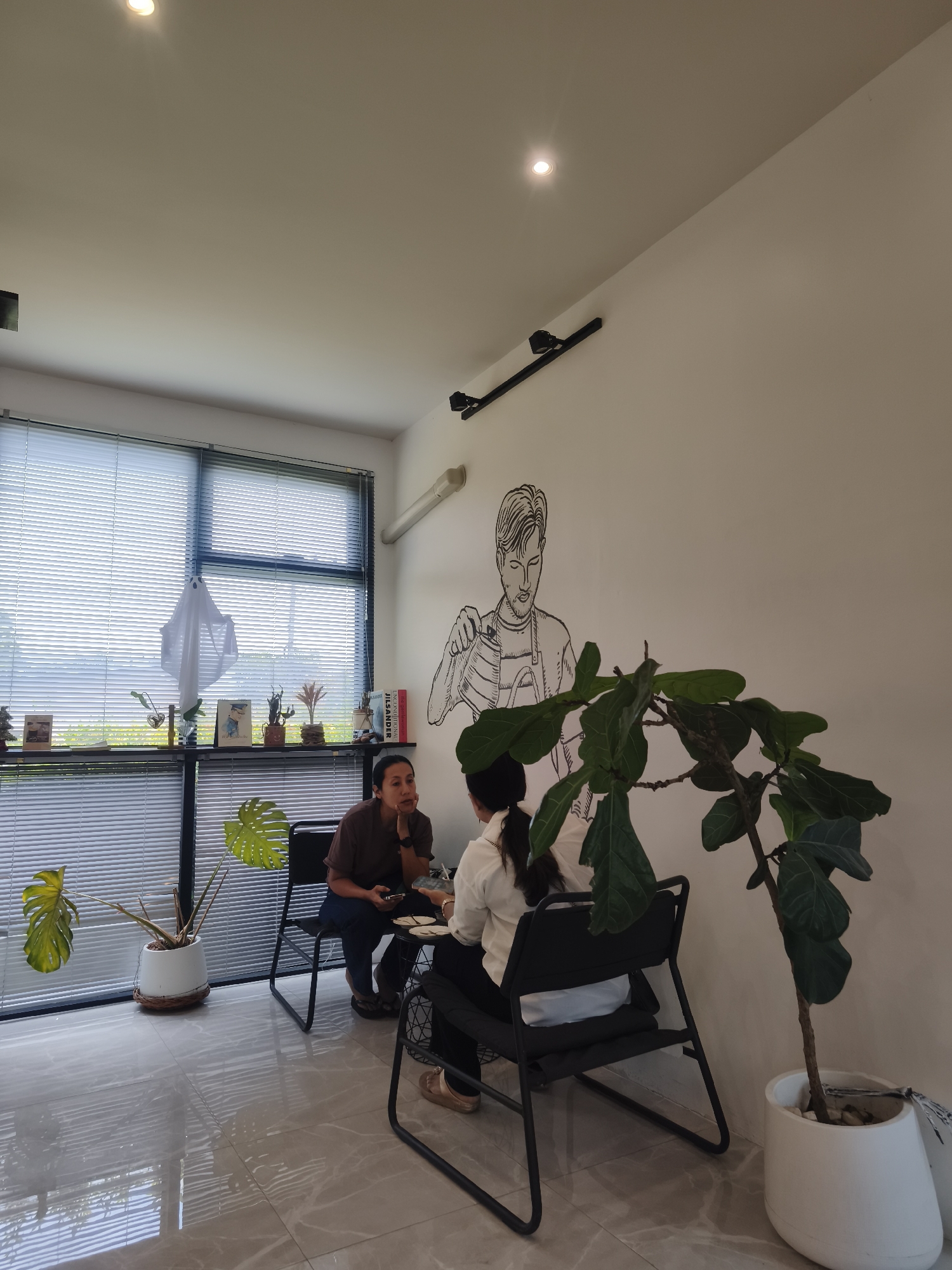 The image shows two people sitting at a table in a modern cafe, possibly located in the city of Ko Samui District, Thailand. The cafe has large windows with white blinds, a white wall with a sketched illustration of a man, and a large potted plant. The floor is tiled and reflective.
One person, wearing a brown t-shirt and dark pants, is sitting on the left, facing the other person. They are holding a phone and appear to be in conversation. The second person, wearing a white shirt and black pants, is seated on the right, facing away from the camera. They are leaning forward, also engaged in the conversation.
On a shelf behind the seated person on the left are various decorative items, including small plants, books, and a white ghost-shaped decoration hanging from the blinds. A large Monstera plant in a white pot is placed near the window. To the right, another large potted plant with broad green leaves, possibly a fiddle-leaf fig, stands in a white planter. The overall ambiance suggests a relaxed and contemporary cafe setting.