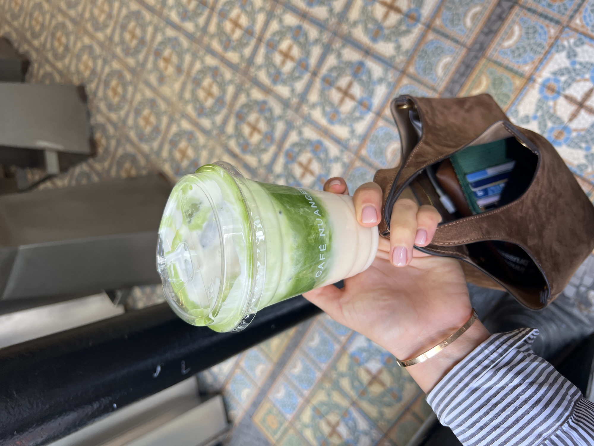 A person is holding a Café Nuancé iced matcha latte in their right hand. A tan suede bag, a gold Cartier bracelet, and a striped button-down shirt are also visible. The location is Paris, France, as implied by the name Café Nuancé on the cup. The background consists of mosaic floor tiles in a pattern of light blues, creams, and tans, suggesting an indoor space, possibly a café or restaurant. A black metallic bar is present on the left side of the image.