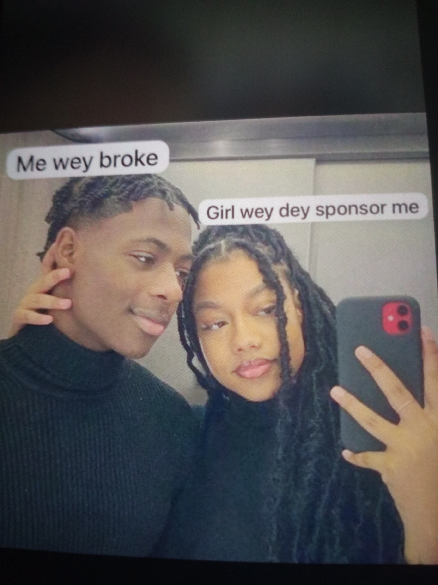 A selfie is taken by a young Black couple in what appears to be an indoor setting with a mirrored surface, possibly an elevator or changing room. The woman on the right is holding a smartphone, angled to capture both herself and the man beside her. The man on the left has a hand gently touching his face, and both individuals are looking towards the camera. They are dressed in dark, high-necked tops. Text overlays are present on the image: "Me wey broke" appears above the man's head, and "Girl wey dey sponsor me" is above the woman's head. The background is plain and metallic. The image does not provide any specific clues about the location within Jalingo, Nigeria.