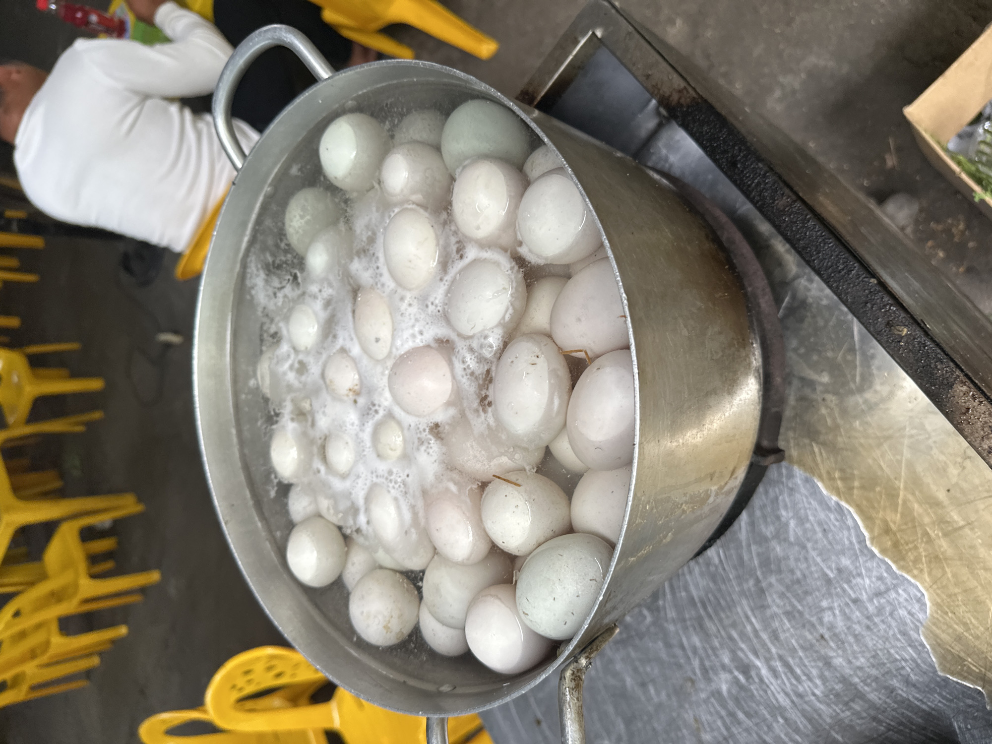 Overhead shot in Nha Trang Ward, Vietnam, shows a large pot filled with numerous eggs submerged in boiling water, positioned on a metallic surface. A person wearing a white long-sleeved shirt is partially visible to the left, holding a red bottle. Stacked yellow chairs are beside the person. The pot, which appears to be made of metal, has two handles and is filled to the brim with eggs. To the right of the pot, a section of a dirty, rusted metal countertop is visible, with a cardboard box partially in view near the edge.