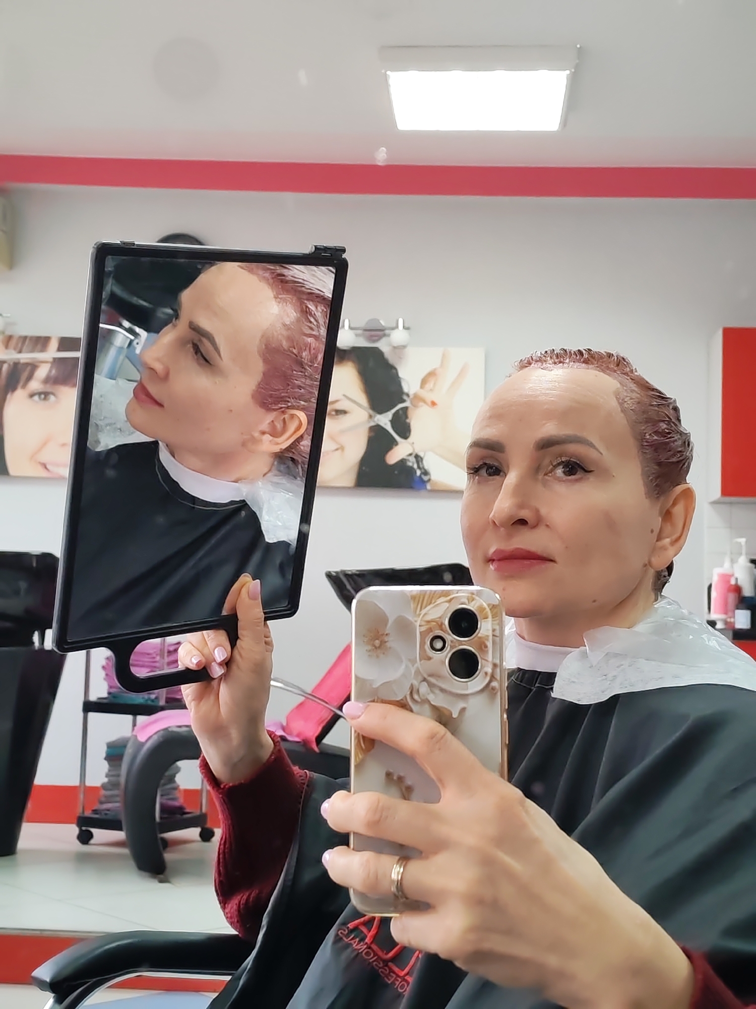 The image depicts a woman taking a selfie in what appears to be a hair salon. The woman has reddish-brown hair that seems to be undergoing a coloring process. She is wearing a black cape over a maroon sweater, and a plastic protective covering around her neck. In her left hand, she holds a rectangular mirror reflecting her profile. Her right hand holds a smartphone with a light-colored case featuring a floral design. A gold wedding band is visible on her left ring finger.
The background of the image shows elements of a hair salon: a black sink station, a rolling cart with items, and framed pictures on the wall depicting women's faces. Red and white paint is visible on the walls and other fixtures, giving the space a modern look. The environment indicates that the photo was taken indoors, in a professional hair care setting. The location of the city of Chaikovsky, Russia is provided.