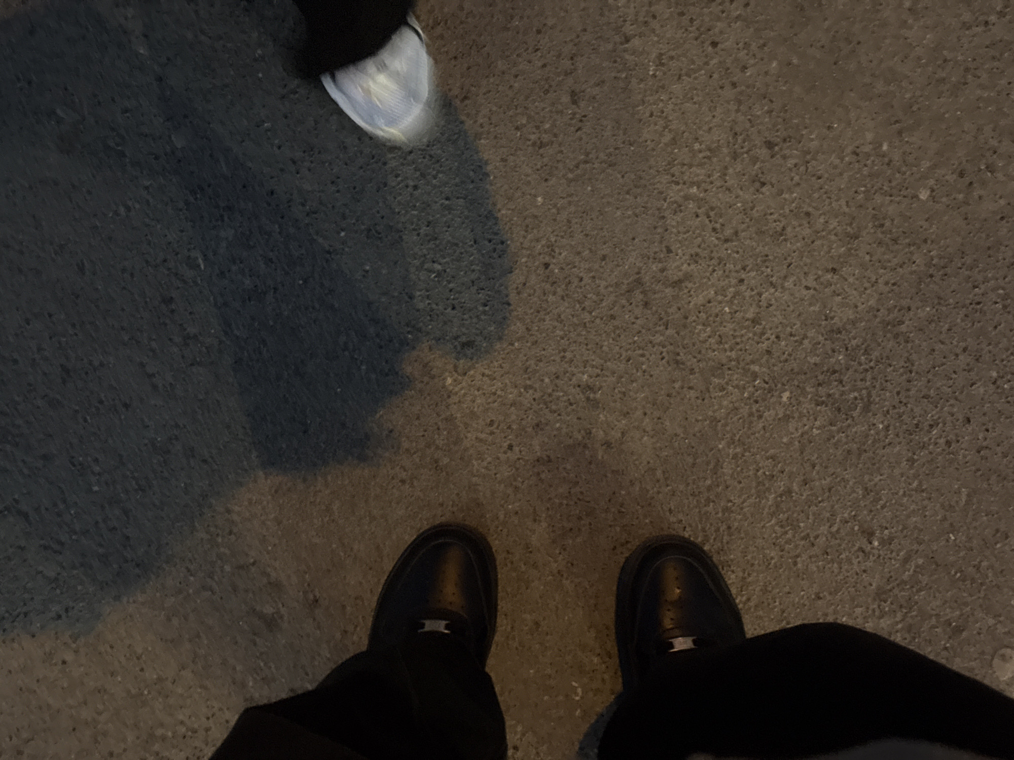 The image, taken in Perm, Russia, shows a low-angle, first-person perspective of two individuals' feet on an asphalt or concrete surface. In the foreground, a pair of black shoes, likely belonging to the photographer, are visible. These shoes appear to be a loafer or dress shoe style, with a distinct, shiny, possibly metallic, rectangular ornament positioned on the top of each shoe. The texture of the shoes suggests leather or a synthetic material.
In the upper left portion of the frame, partially visible, is the front of a white or light-colored athletic shoe, indicating the presence of another person standing nearby.
The ground surface is dark, granular, and appears to be made of asphalt or rough concrete. A large, dark shadow casts across the left side of the frame, suggesting a light source, possibly artificial street lighting, is positioned to the right of the scene. The overall lighting is dim, indicating it is likely nighttime or dusk. No specific activities beyond standing or walking are discernible, and no text is visible in the image.
