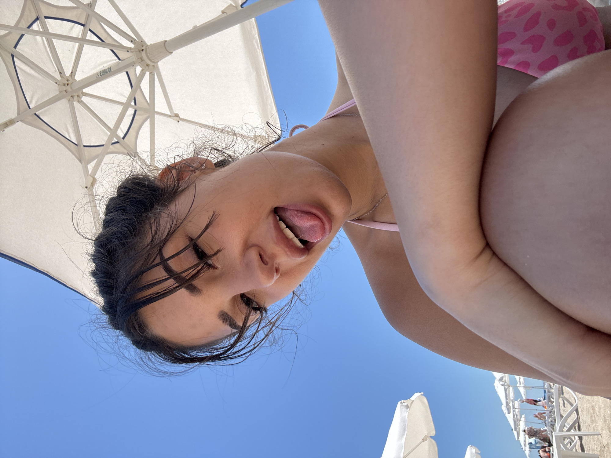 A young woman with dark hair pulled into a bun is photographed from a low angle on a sunny beach, likely in Sozopol, Bulgaria, given the context of the image. She is wearing a pink bikini with a heart pattern. Her face is tilted towards the camera, with her tongue sticking out. Her right arm is bent and resting across her chest, obscuring part of her body. Behind her, a white beach umbrella is visible, along with a bright blue sky. Further in the background, beach chairs and other umbrellas suggest a populated beach area. The lighting indicates it is daytime, and the overall scene conveys a casual, playful mood.