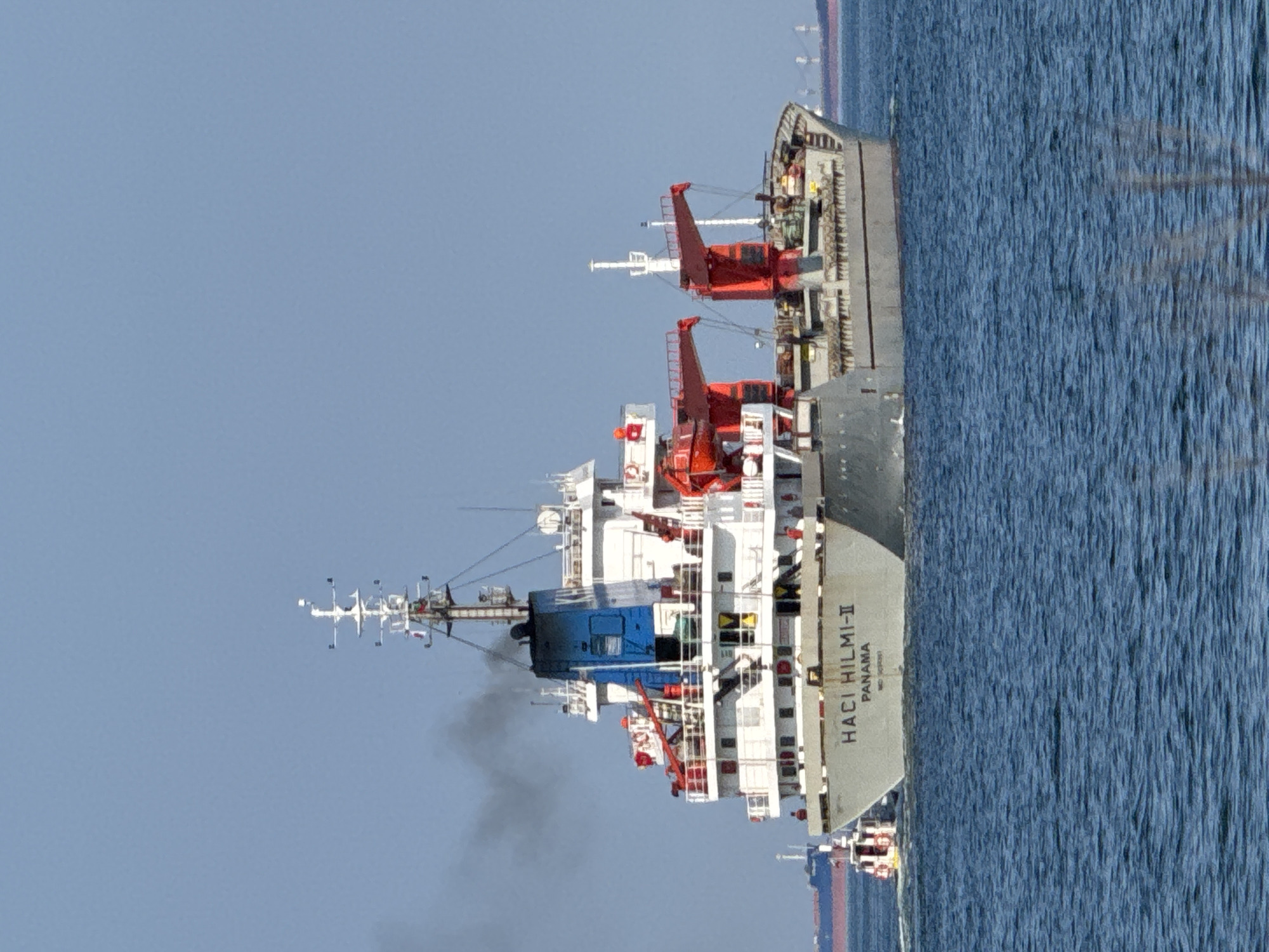 The image shows a cargo ship sailing on the water, likely the Black Sea near Varna, Bulgaria. The ship's name, "HACI HILMI-II PANAMA," is visible. The ship is white and gray with red cranes. Black smoke is emanating from a portion of the ship. The sky is light blue and the water is a darker blue.