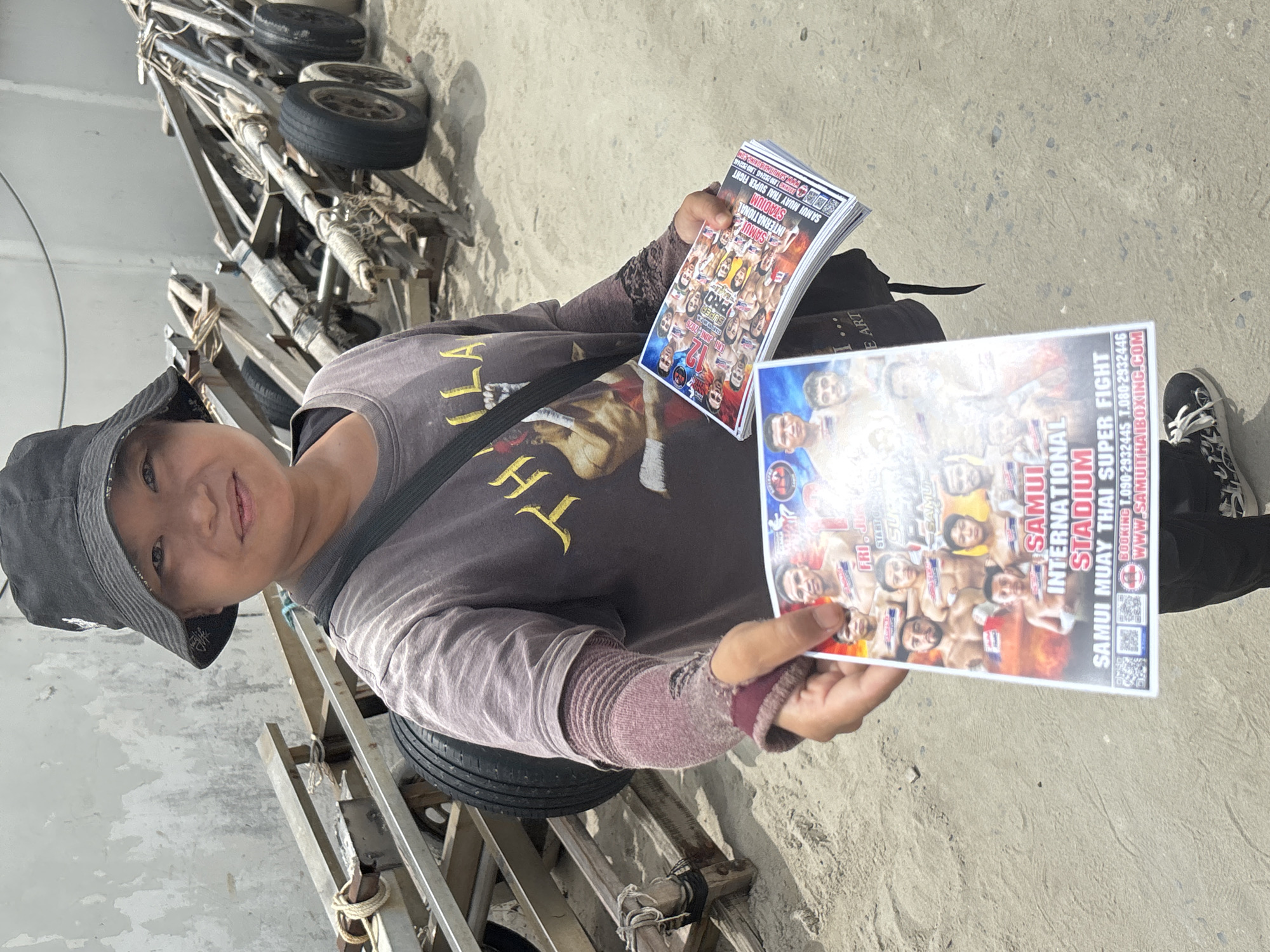 A person wearing a black bucket hat and a grey t-shirt with a graphic on it stands outdoors on a sandy surface. They are holding two stacks of flyers promoting a Muay Thai boxing event. The flyers feature images of fighters and event details. In the background, a boat trailer with multiple wheels and wooden supports is visible, along with a weathered concrete wall. The scene appears to be in Baan Chaweng, Thailand, given the promotional material for "Samui International Stadium" and "Samui Muay Thai Super Fight".
