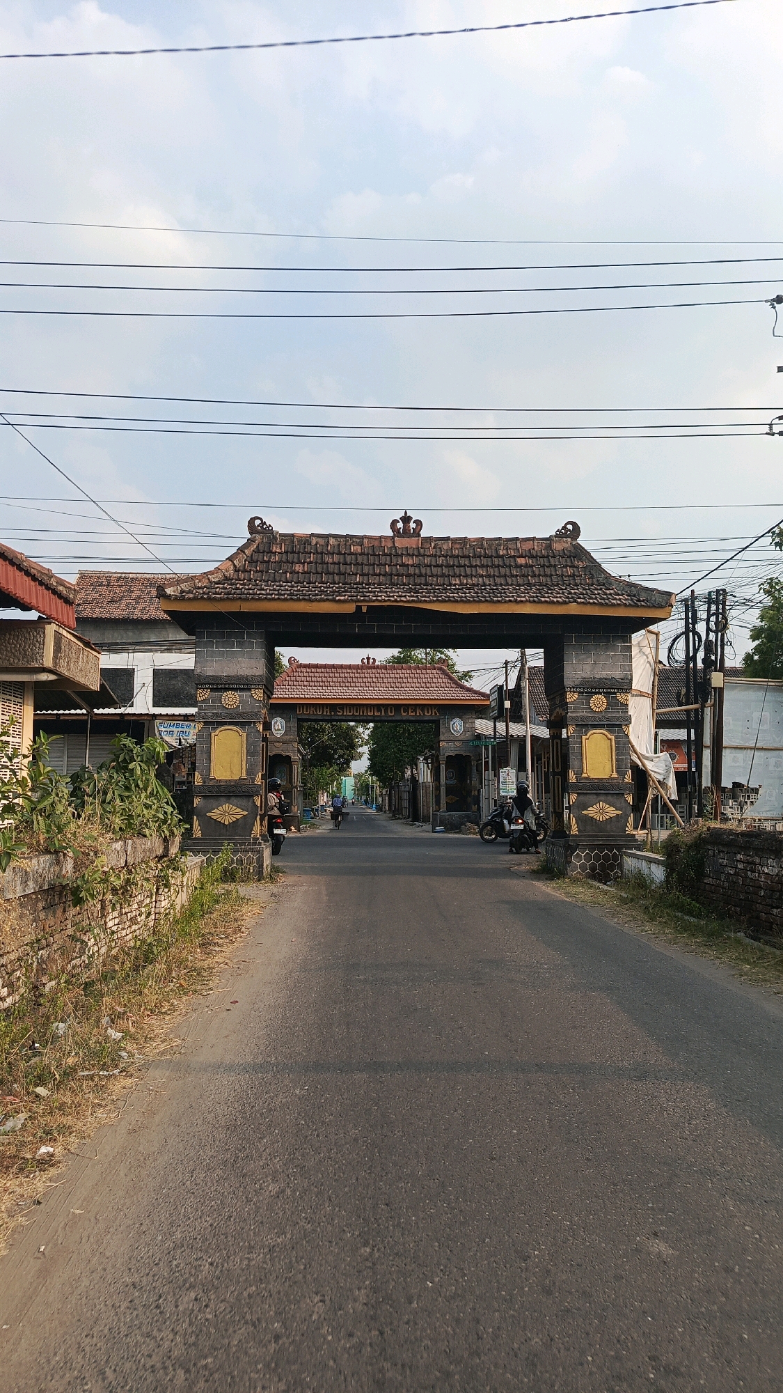 This is a forward-facing, eye-level shot of a gateway arch in Patihan Wetan, Indonesia. The arch is constructed of dark stone with decorative gold accents and a tiled roof. The words "Dukuh.Sido Mulya.Cekok" are visible on the arch. The arch spans a paved road. On the left side of the road, there is a low brick wall with dry grass and some debris. On the right side, there are utility poles and wires overhead. A few motorcycles and a bicycle are present on the road, with some people visible. The sky is partly cloudy. Buildings are visible on either side of the road beyond the arch.