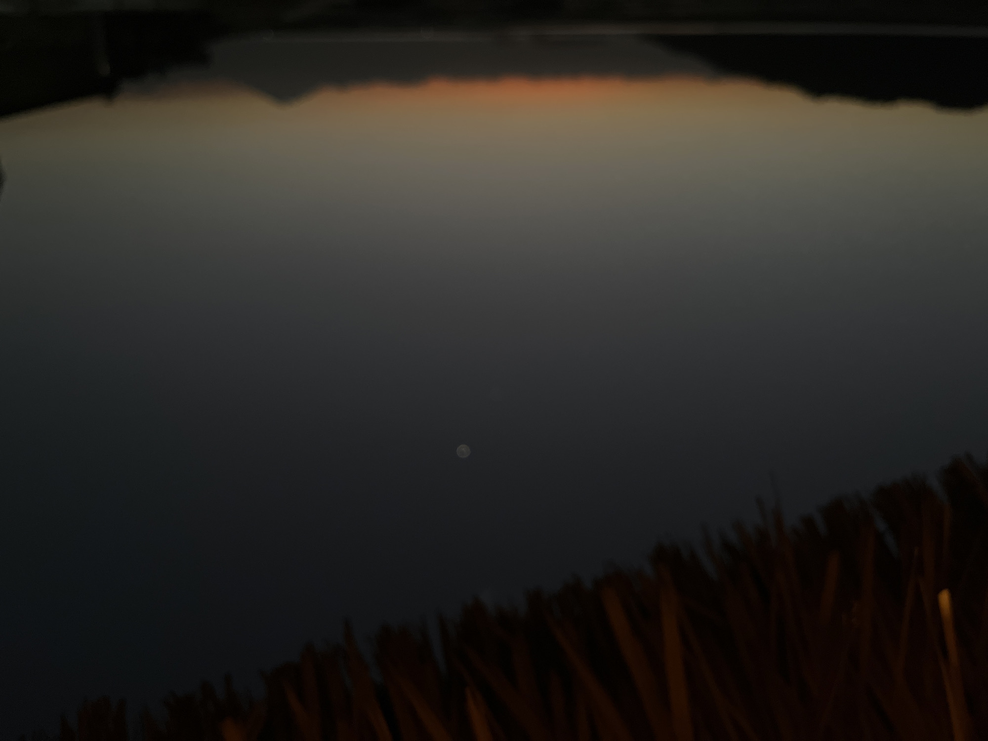 The image captures a twilight scene over a body of water, likely the ocean given the coastal silhouette of landmasses. The sky transitions from a dark, dusky blue near the top to lighter shades of gray and orange near the horizon, indicating either sunset or sunrise. A blurred, distant mountain range is visible against the horizon. In the foreground, the edge of a thatched roof or awning, made of dried plant material, partially obscures the top portion of the image. A small, circular, bright object, possibly the moon or a bright star, is visible in the upper-middle section of the dark sky. The overall scene is dark and slightly out of focus, creating a moody atmosphere. The location is Kertasari, Indonesia.