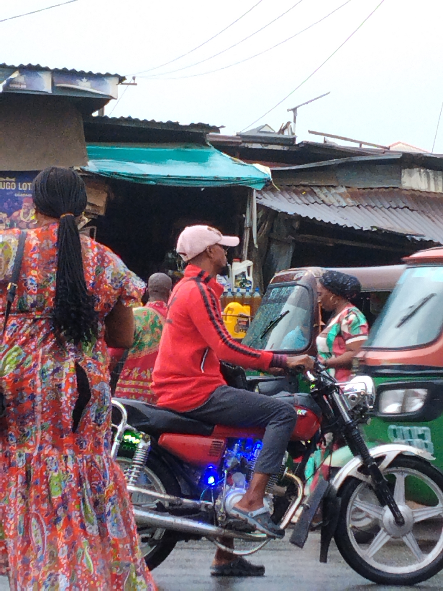 The image shows a street scene in Aba, Nigeria. A man is sitting on a red motorcycle, wearing a pink cap, red jacket, and gray pants. He is wearing light-colored sandals. Blue LED lights are visible underneath the motorcycle seat. A woman wearing a colorful orange, red, and blue dress stands in the foreground. Another woman wearing a patterned headscarf and dress is standing by a parked auto-rickshaw (tricycle) with a green exterior. Several other people are visible in the background. Oil bottles are stacked behind the motorcycle and next to the first auto-rickshaw. Overhead, corrugated metal roofing is visible.
