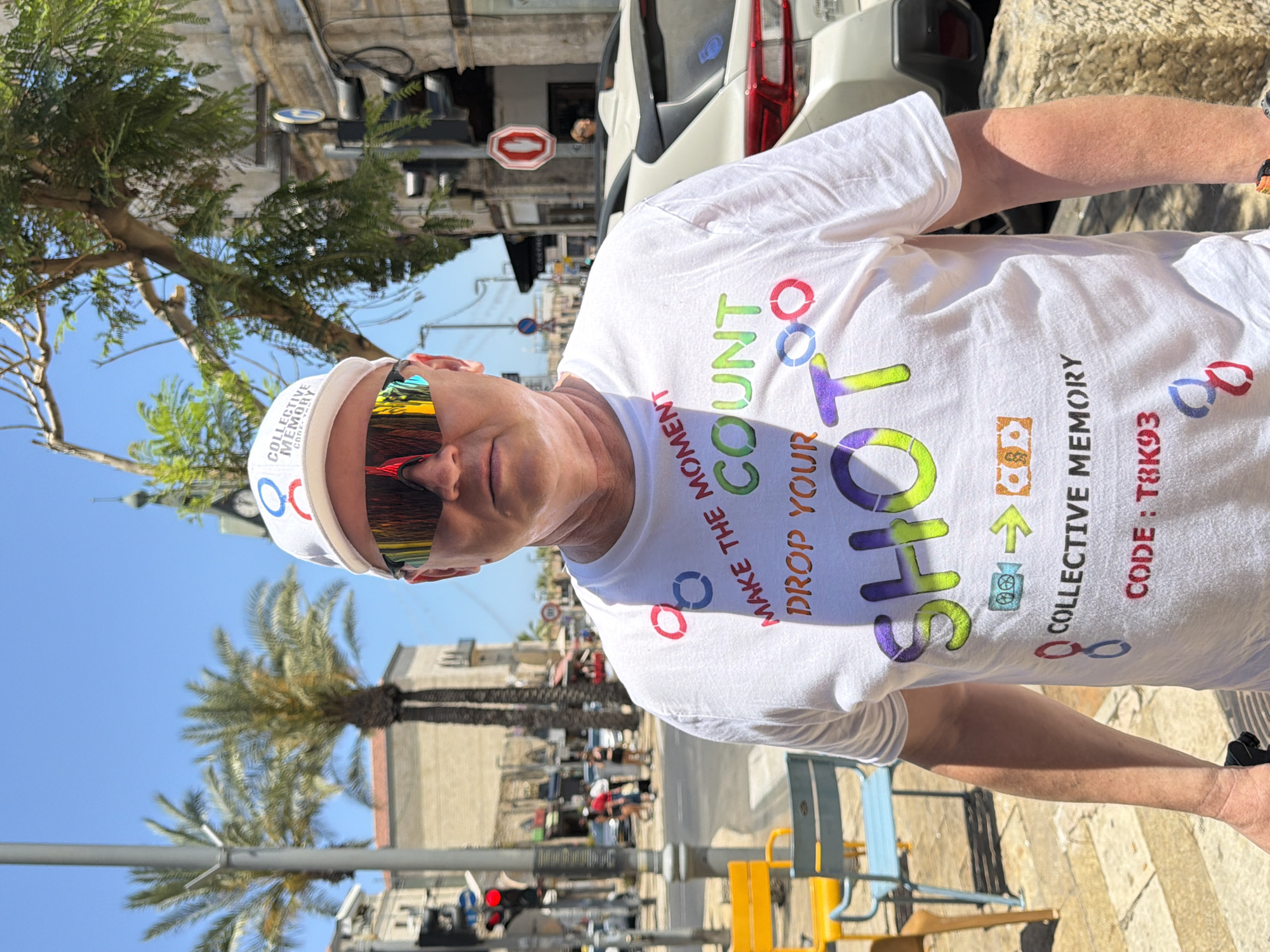 A medium-shot photograph depicts a fair-skinned man wearing mirrored sunglasses and a white hat with circular designs, likely standing in Tel Aviv, Israel, as indicated by the Hebrew word "תל-אביב" on his hat. He is wearing a white t-shirt adorned with the words "MAKE THE MOMENT COUNT," "DROP YOUR SHOT," and "COLLECTIVE MEMORY CODE: T8K93" along with colorful circular designs and camera icons. The background reveals an urban landscape featuring palm trees, buildings, and a street with a parked white car and pedestrians. A street sign with a stop symbol is visible in the background. The lighting suggests a bright, sunny day.