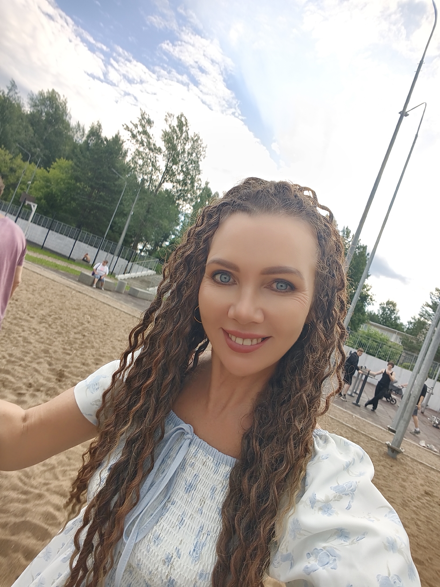 A woman with long, curly brown hair and blue eyes is taking a selfie outdoors. She is wearing a white dress with a blue floral pattern. She is smiling and looking at the camera. The background appears to be a park or outdoor recreation area, with sand on the ground, trees and buildings in the distance, and metal poles that may be part of a playground or sports equipment. Several other people are visible in the background, some of whom appear to be exercising. The sky is cloudy with patches of blue. The scene is likely in the city of Chaikovsky, Russia, as indicated by the presence of Russian text on some of the structures in the background (not clearly legible in this resolution).