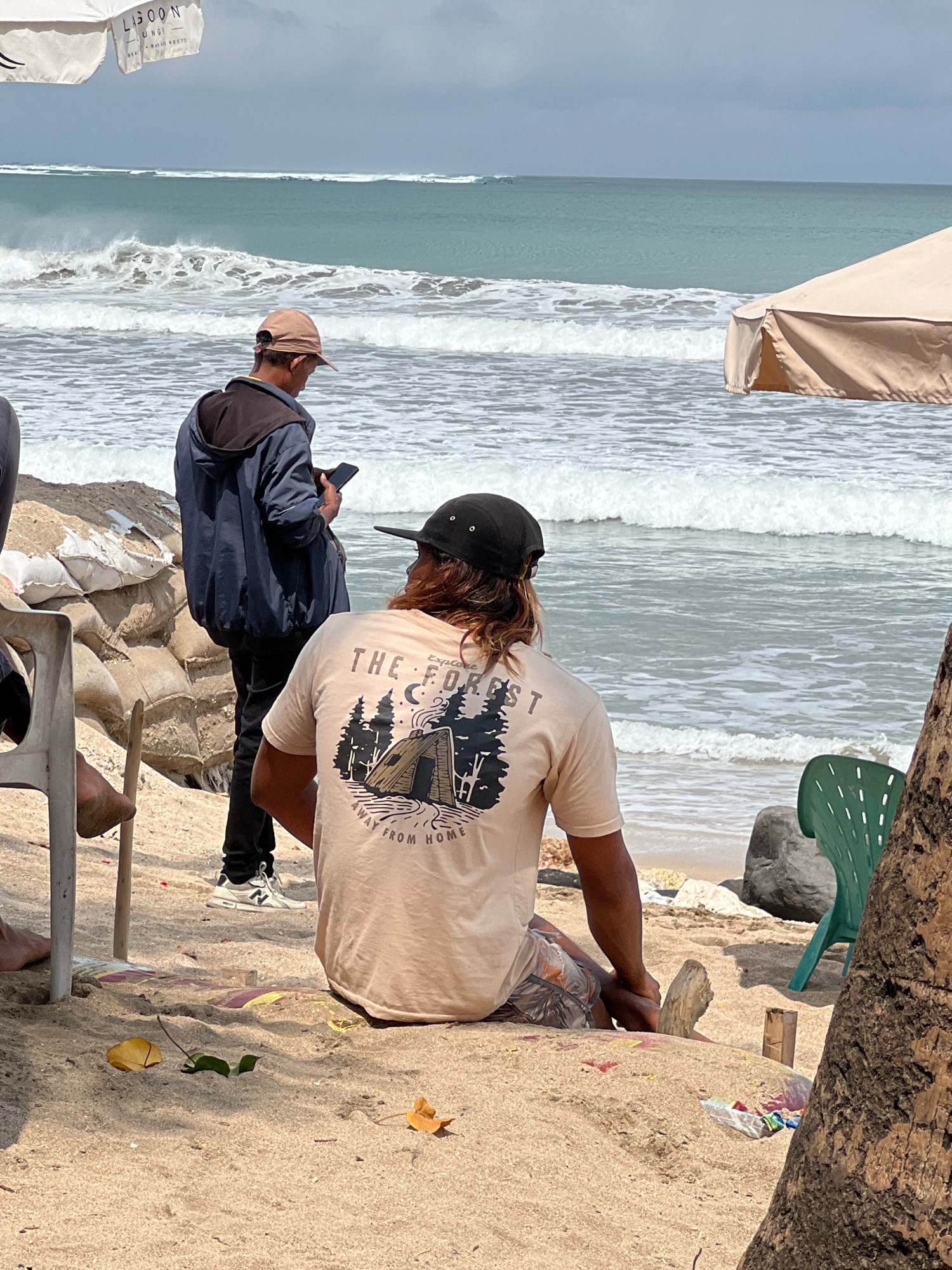 The image shows a beach scene in Kuta, Indonesia, on a cloudy day. In the foreground, a person with long, brown hair, wearing a tan t-shirt with a graphic print and patterned shorts, sits on the sand with their back to the camera. They are facing the ocean, which has moderate waves. To the left and slightly behind them, another person stands wearing a dark jacket, black pants, and a light brown baseball cap, looking at their phone. Sandbags are piled up behind this person, and a white umbrella with text is visible in the upper left corner, suggesting a beach lounge or establishment. A light-colored tent or canopy is visible in the middle ground to the right. The beach is sandy with some debris on the ground, including leaves and plastic bags. A green plastic chair is positioned on the sand to the right of the frame, partially obscured by a tree trunk. The ocean is a turquoise blue, and the sky is overcast.